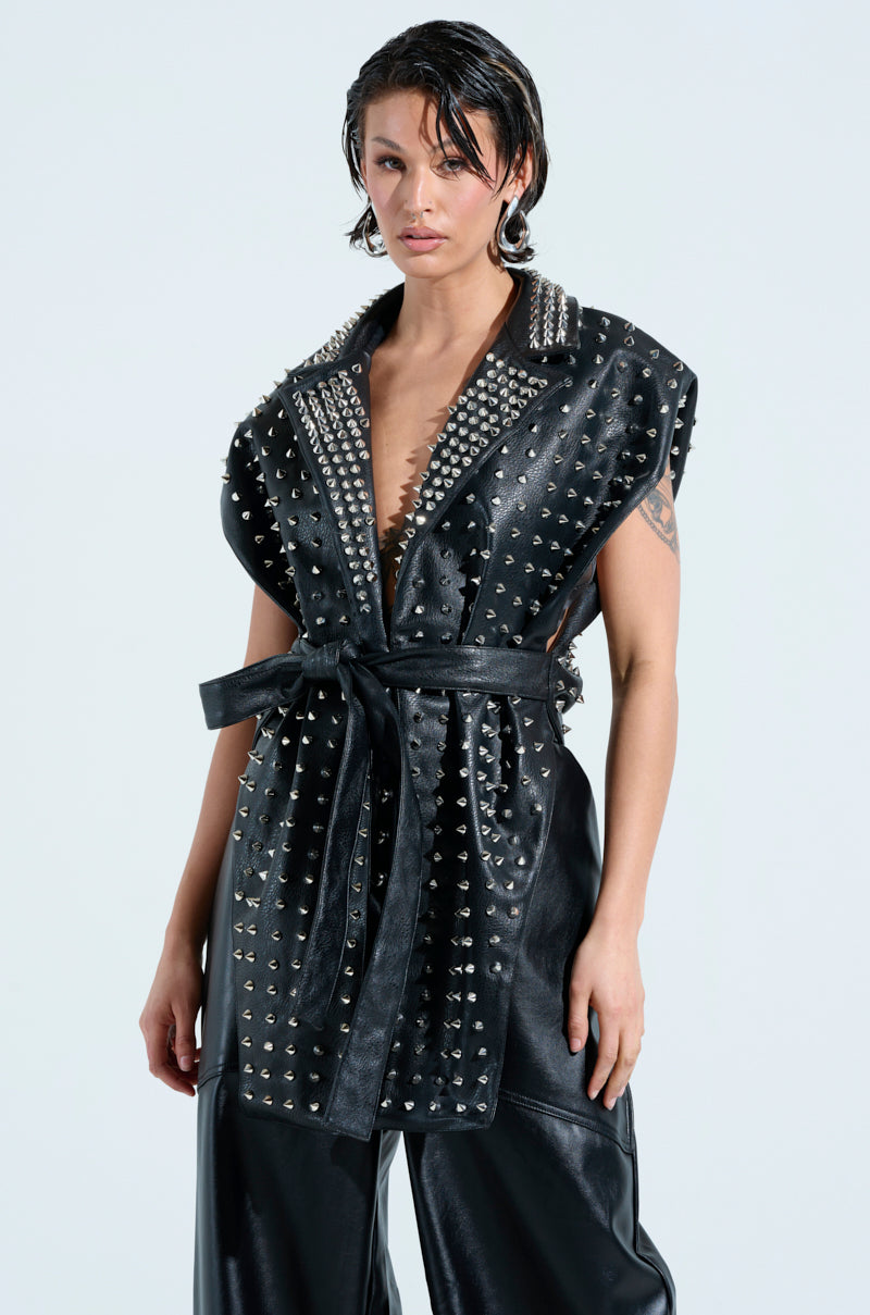 A person confidently poses in the TELL ME ABOUT IT STUDDED VEST, a sleeveless black leather piece with striking spike studs, complemented by sleek black leather pants. Their short, wet-look hair perfectly matches the edgy faux leather ensemble against a plain background.