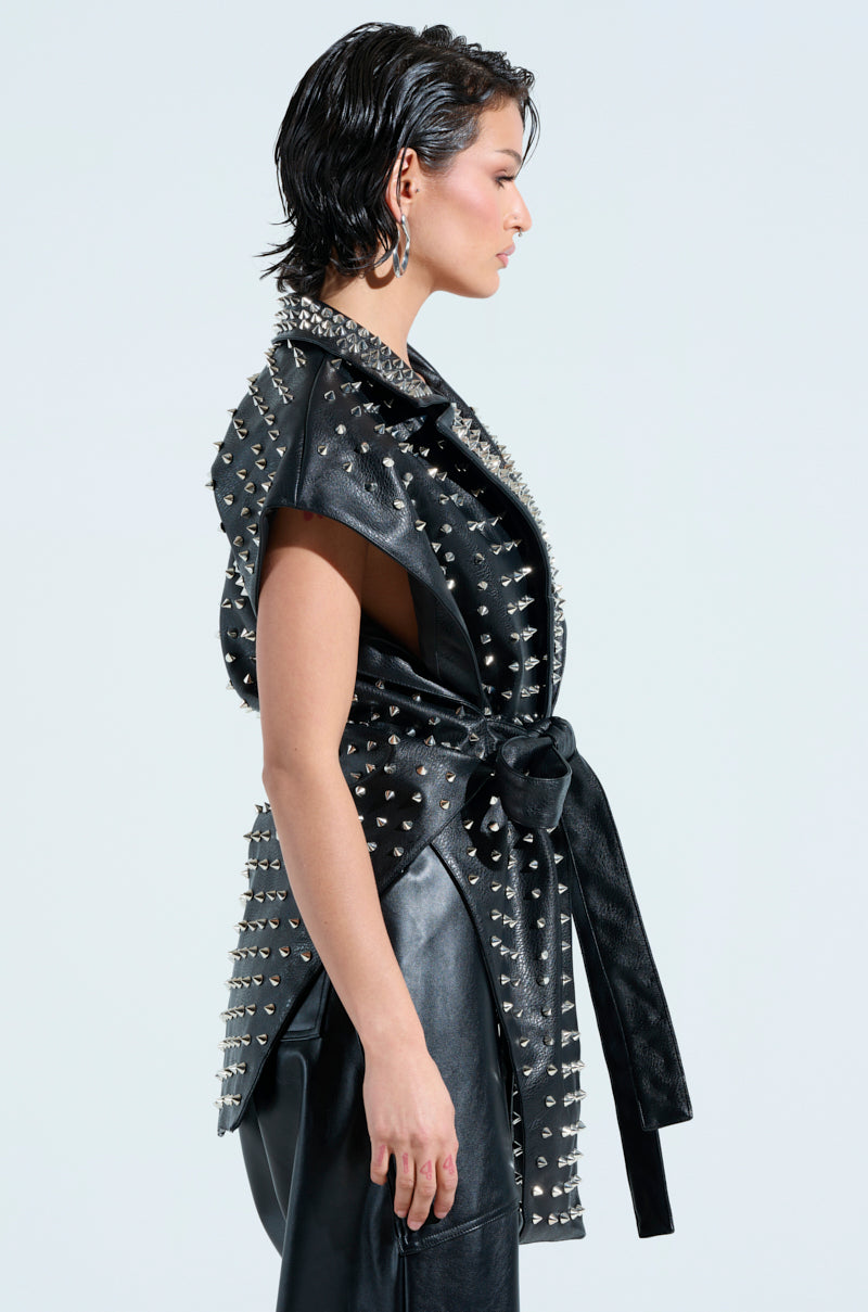 Wearing the TELL ME ABOUT IT STUDDED VEST, a person with short, wet-look hair dons a black, sleeveless faux leather jacket adorned with spike studs and matching leather pants while standing in profile against a light background.