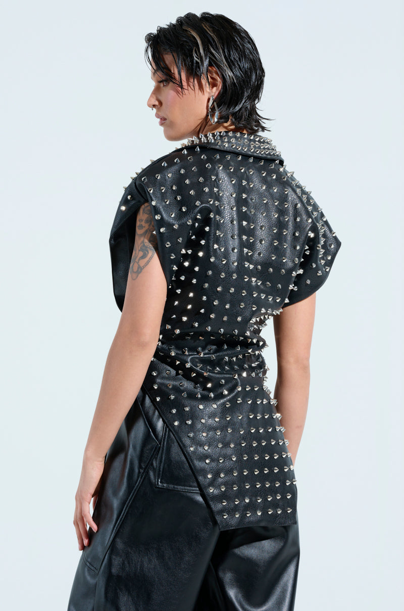 A person with short dark hair and visible tattoos wears the TELL ME ABOUT IT STUDDED VEST in black, paired with faux leather pants. Their punk-inspired look is completed with hoop earrings and spike stud embellishments, all set against a plain white backdrop.
