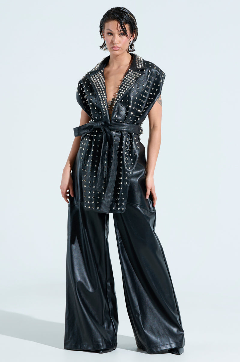 A person is wearing the TELL ME ABOUT IT STUDDED VEST, a sleeveless black faux leather vest with spike stud embellishments, over wide-leg pants. The vest is tied at the waist with a sash. They have a short, slicked-back hairstyle against a plain background.