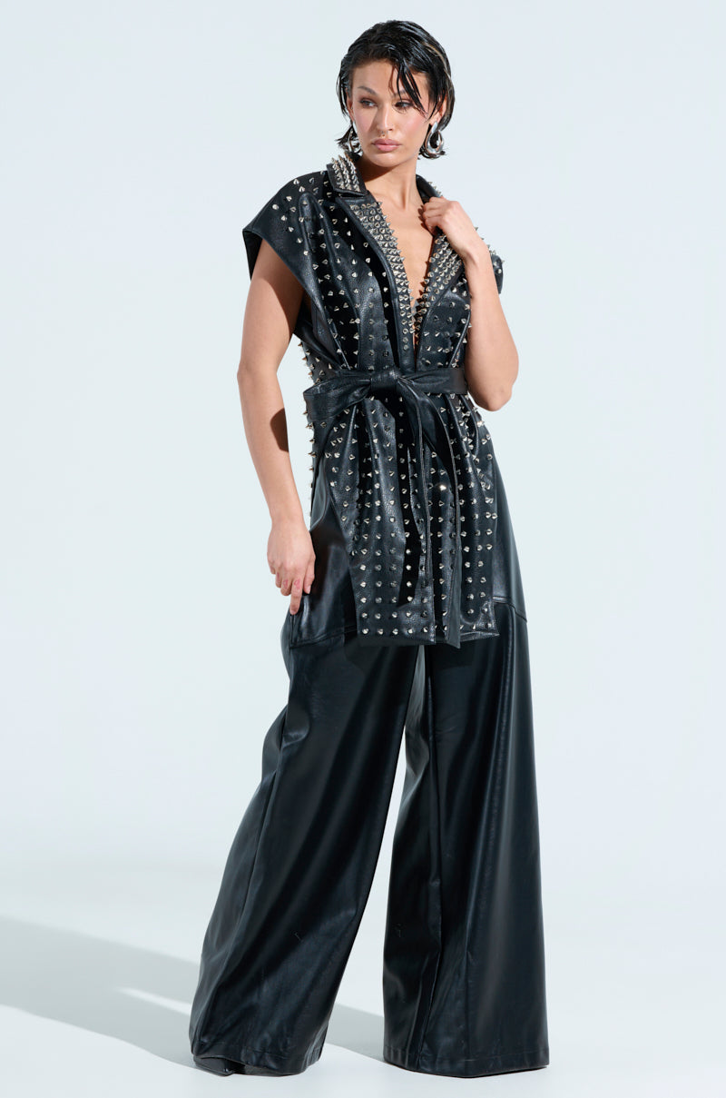 A woman confidently poses in a black TELL ME ABOUT IT STUDDED VEST made of faux leather, with spike studs, a deep V-neck, and wide-legged pants. Her hair is styled wet, and she accessorizes with statement earrings against a plain light background.