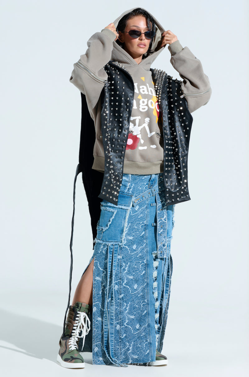 A person confidently adjusts their sunglasses while wearing the TELL ME ABOUT IT STUDDED VEST with spike stud embellishments over a gray hoodie with colorful text, paired with layered denim skirts, and completed by green and brown high-top sneakers.