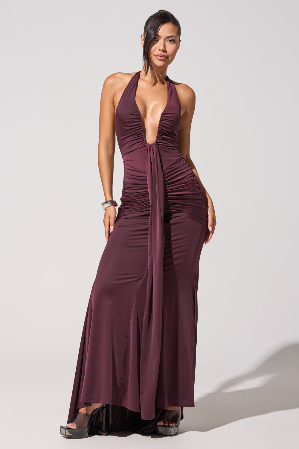 A woman stands confidently against a plain background wearing the SET THE MOOD MESH HALTER MAXI DRESS in purple, featuring a plunge neckline and ruched detailing. She pairs it with platform shoes and minimal jewelry. 2