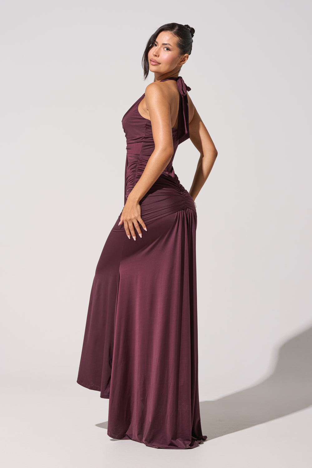 A woman poses sideways in the SET THE MOOD MESH HALTER MAXI DRESS—a sleeveless, long burgundy gown with a halter neckline and side slit—against a plain light gray background. 5