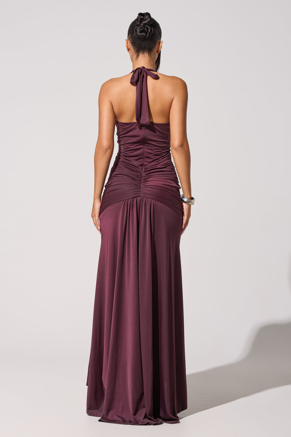 A woman stands facing away in the SET THE MOOD MESH HALTER MAXI DRESS—a floor-length, sleeveless purple maxi with a ruched back. Her hair is styled in a neat bun and she wears a bracelet. The background is plain and light-colored. 6