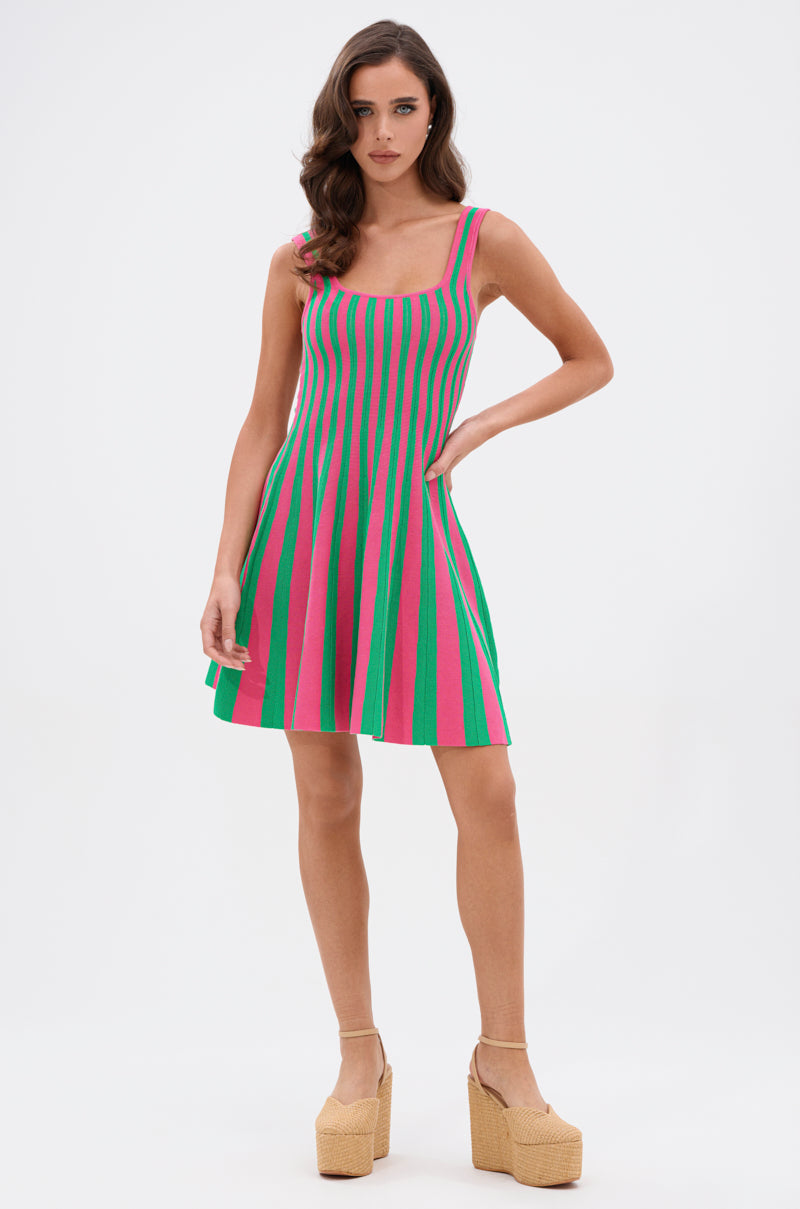 A woman with wavy brown hair wears the GIRLY POP STRIPE MINI DRESS, a sleeveless, ribbed mini dress with vertical green and pink stripes, paired with beige platform heels against a plain white background.