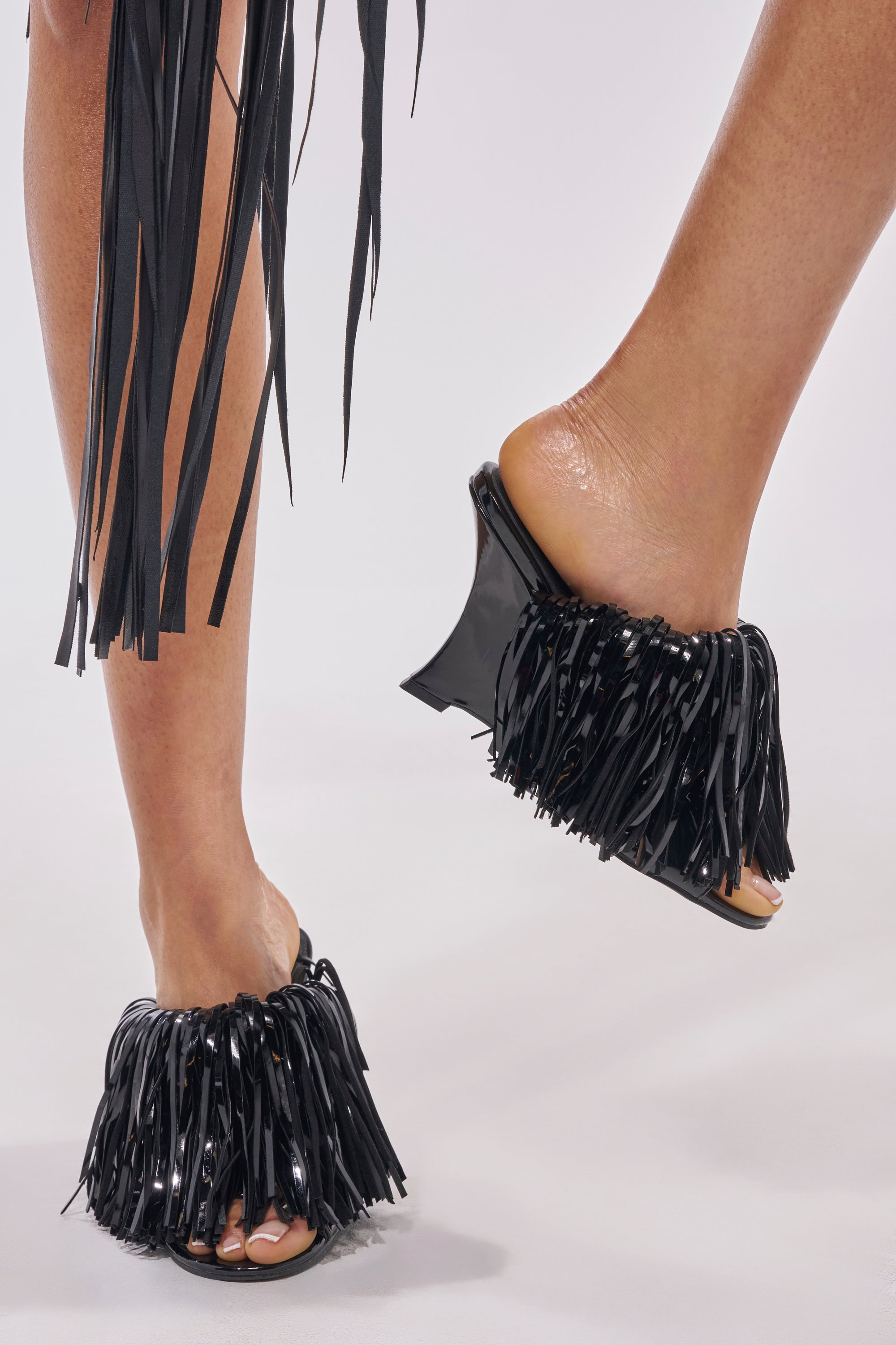 A person wears AZALEA WANG VAILYN BLACK FRINGE PU WEDGE SANDALS featuring black open-toe design and fringe embellishments, with their feet visible against a plain light background. 1