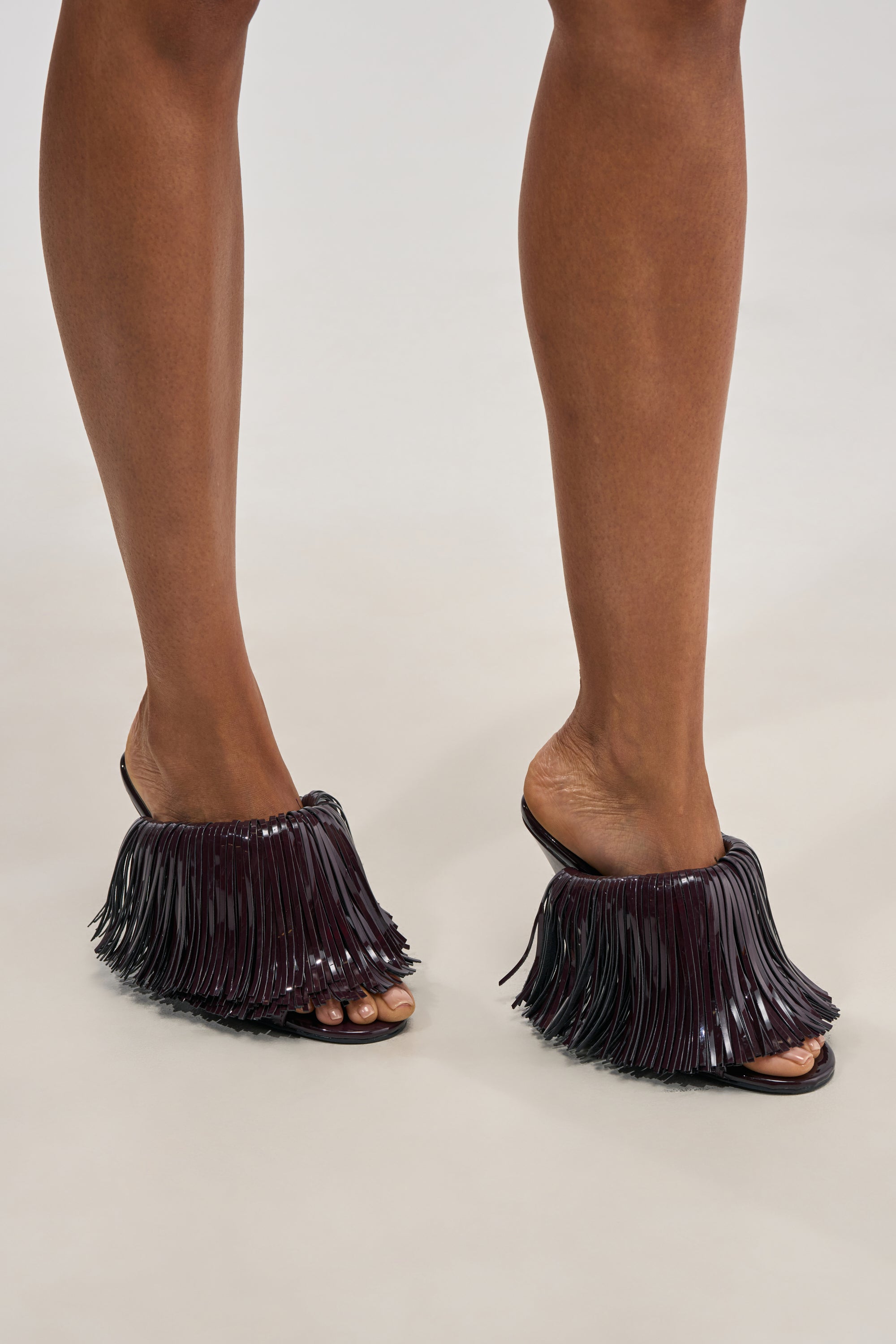 A person wearing the AZALEA WANG VAILYN PURPLE PATENT FRINGE PU WEDGE SANDAL, featuring shiny long fringe details and a bold open-toe wedge design—perfect party footwear—standing on a light-colored surface.