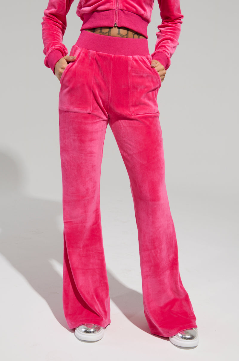A person wears the Velour Galore Straight Leg Jogger in pink, featuring a zip-up jacket and flared pants. Their hands rest in the pockets as they stand against a plain background, with the look finished by silver sneakers.
