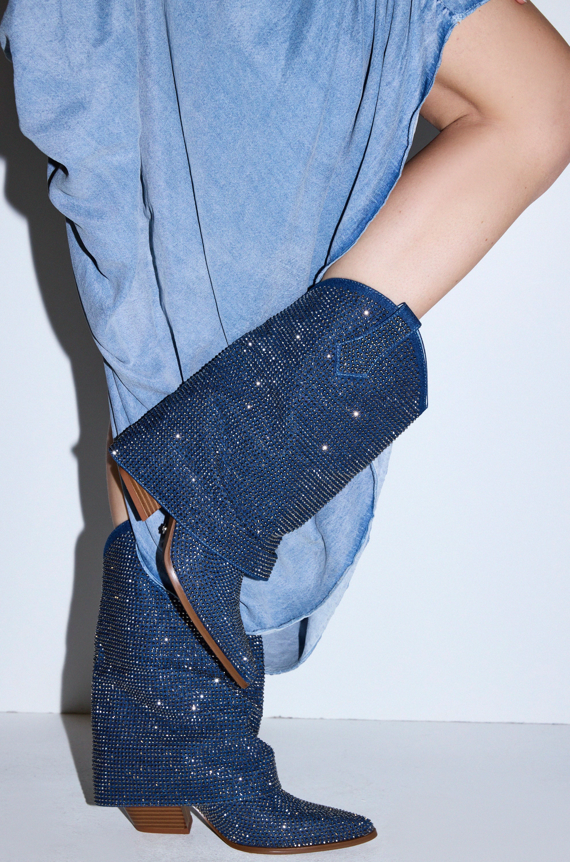 A person in a light blue rolled-up dress poses with one knee raised, featuring the AZALEA WANG VANADIS DENIM RHINESTONE COWBOY BOOTIE—tall denim boots with rhinestones, pointed toes, and low wooden heels—against a plain white background.