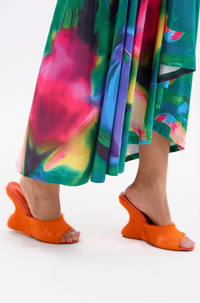 A person wearing a brightly colored, abstract-patterned skirt and the AZALEA WANG VASA ORANGE FURRY WEDGE SANDAL with an open almond toe stands on a white background. 3