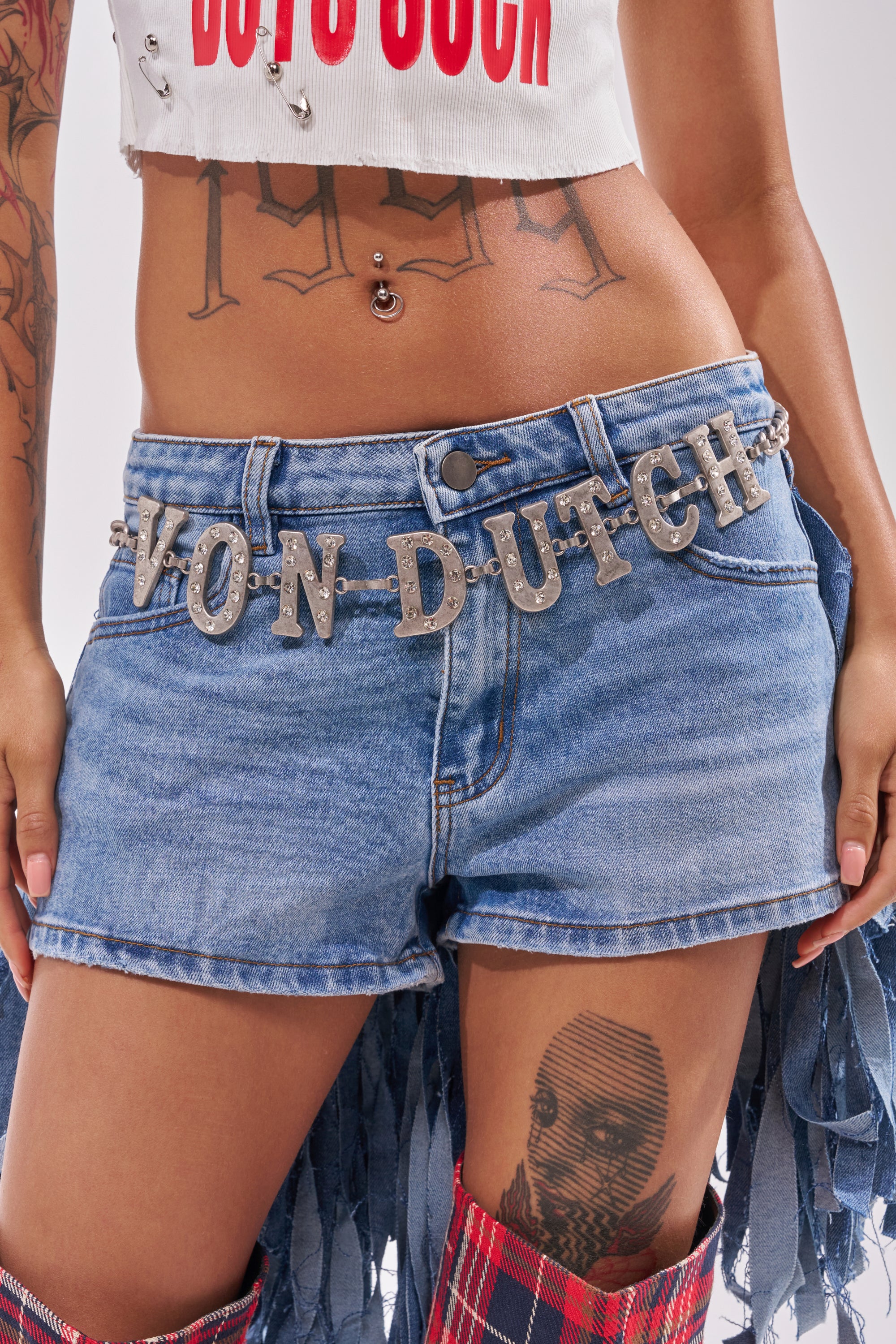 A person wearing denim shorts, a white crop top with red text, tattoos on their torso and thigh, and the VON DUTCH RHINESTONE CHAIN BELT. 0