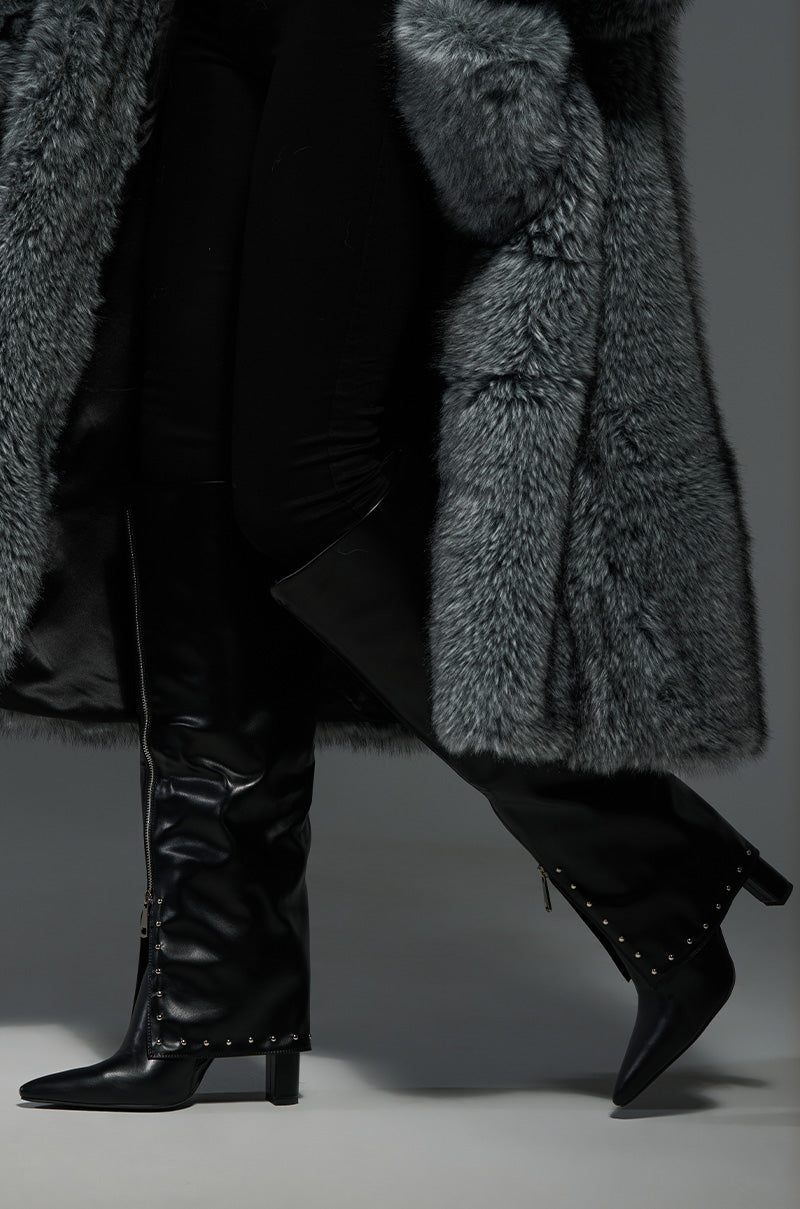 A person wearing a long, fluffy gray fur coat and sleek black knee-high boots, identified as the AZALEA WANG VEEVEE BLACK STILETTO BOOT, with chunky heels and small studs along the soles, standing on a gray background.