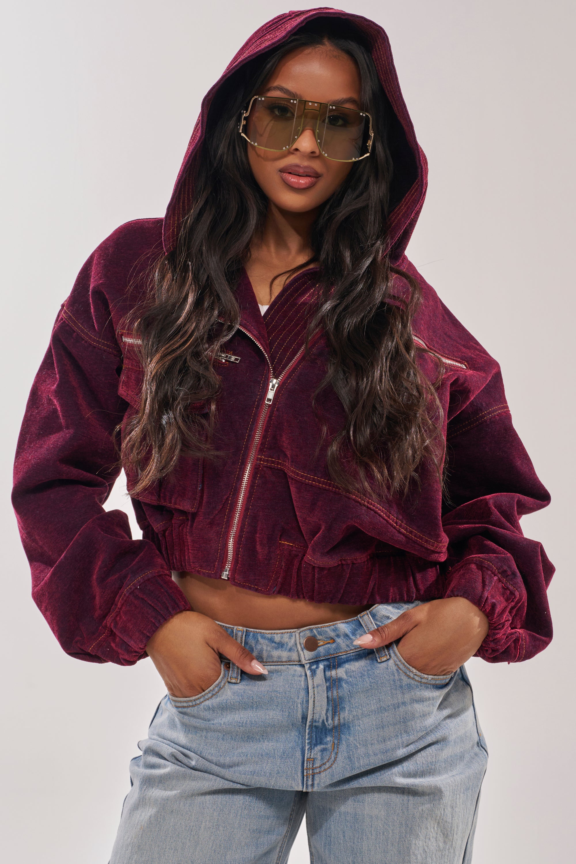 A woman models the PAYTON VELVET DENIM BOMBER—a maroon velvet jacket with an oversized hood—paired with large sunglasses and light blue jeans, hands in pockets against a plain background. 0