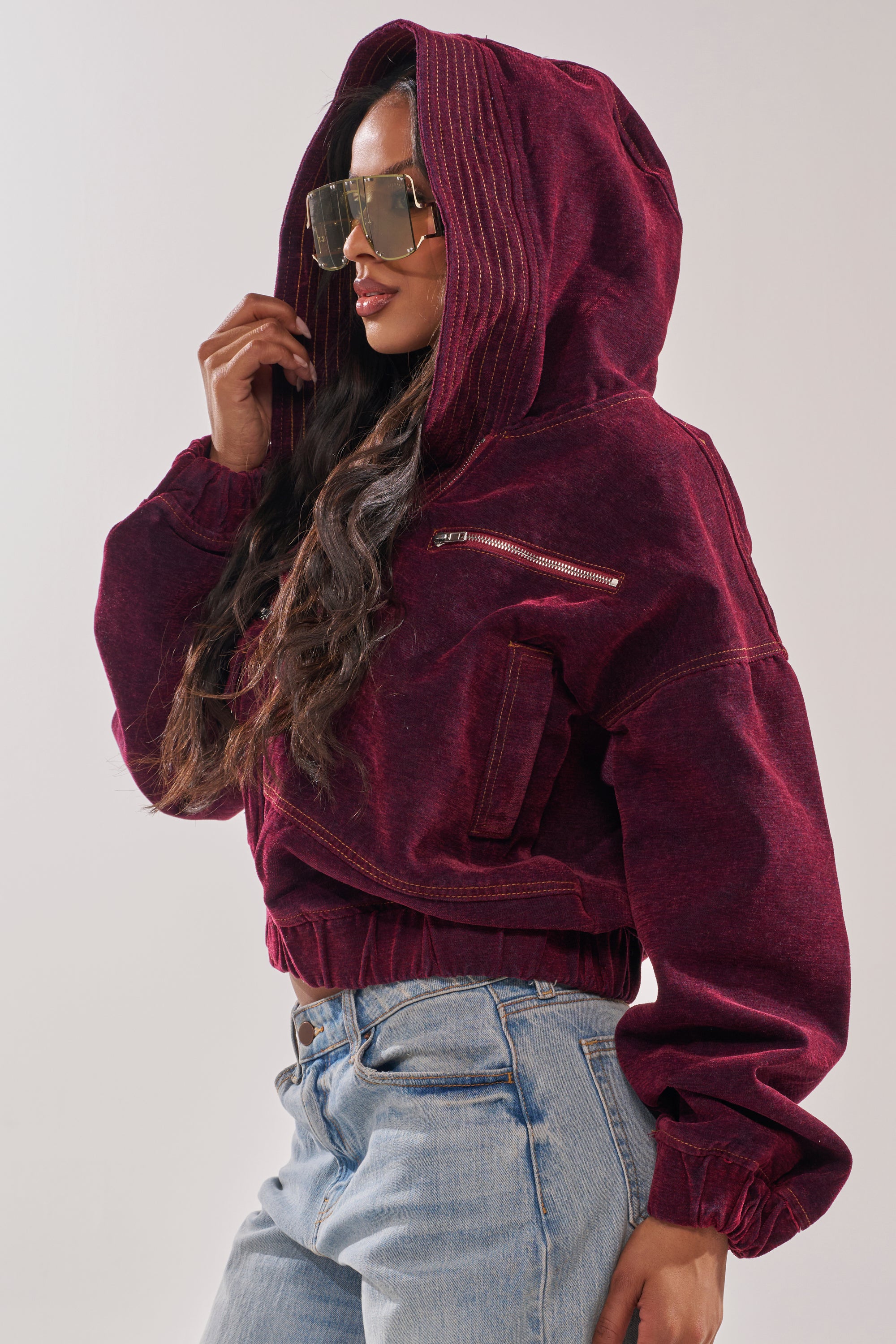 A woman models the PAYTON VELVET DENIM BOMBER, an oversized hooded jacket in burgundy, paired with light blue jeans and large sunglasses, posing sideways against a plain background. 4