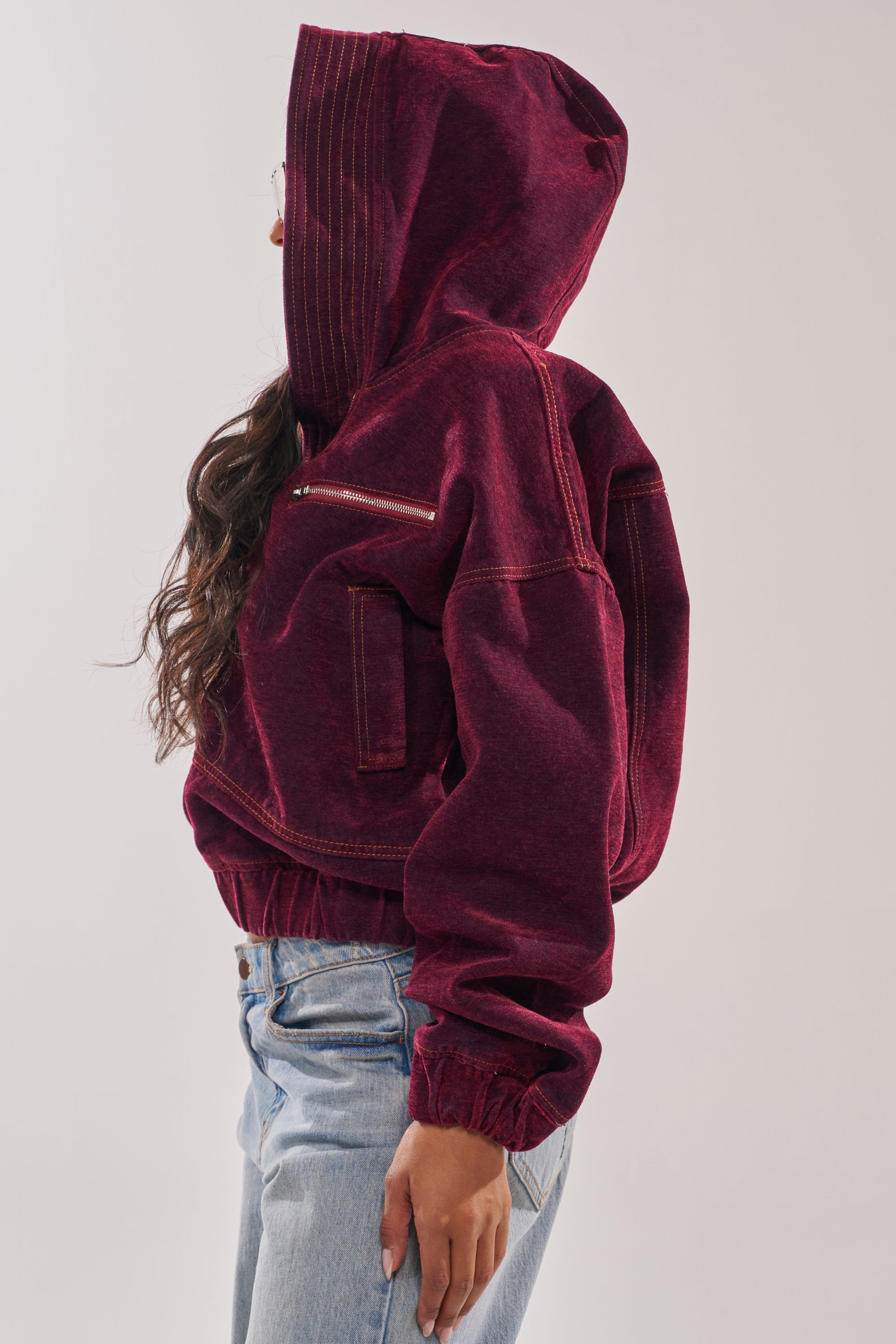 A person in profile wears the PAYTON VELVET DENIM BOMBER—an oversized maroon velvet jacket with white stitching—paired with light blue jeans, standing against a plain light background. 5