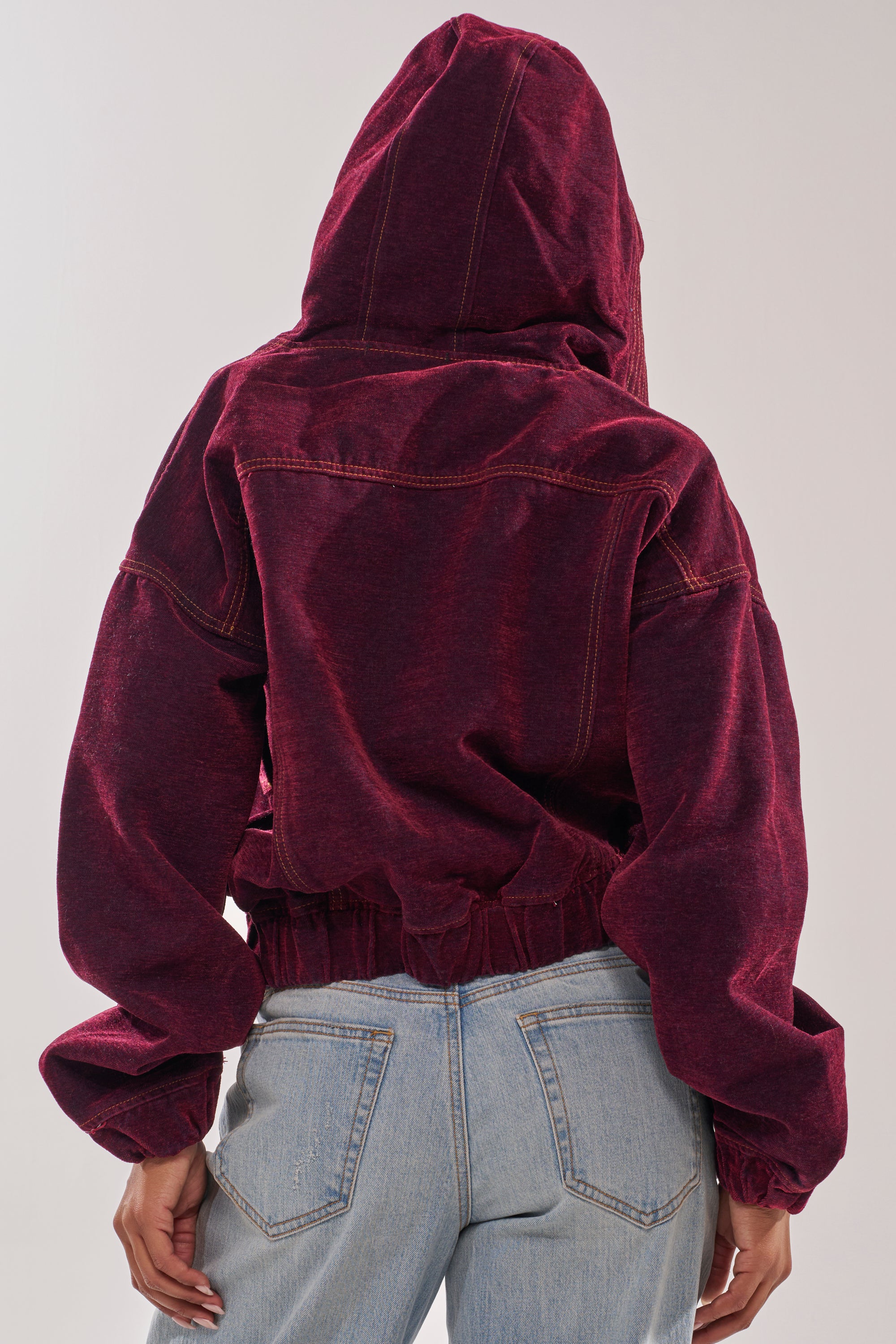 Seen from the back against a neutral background, a person wears the PAYTON VELVET DENIM BOMBER—a maroon velvet jacket with an oversized hood—paired with light blue high-waisted jeans. 6