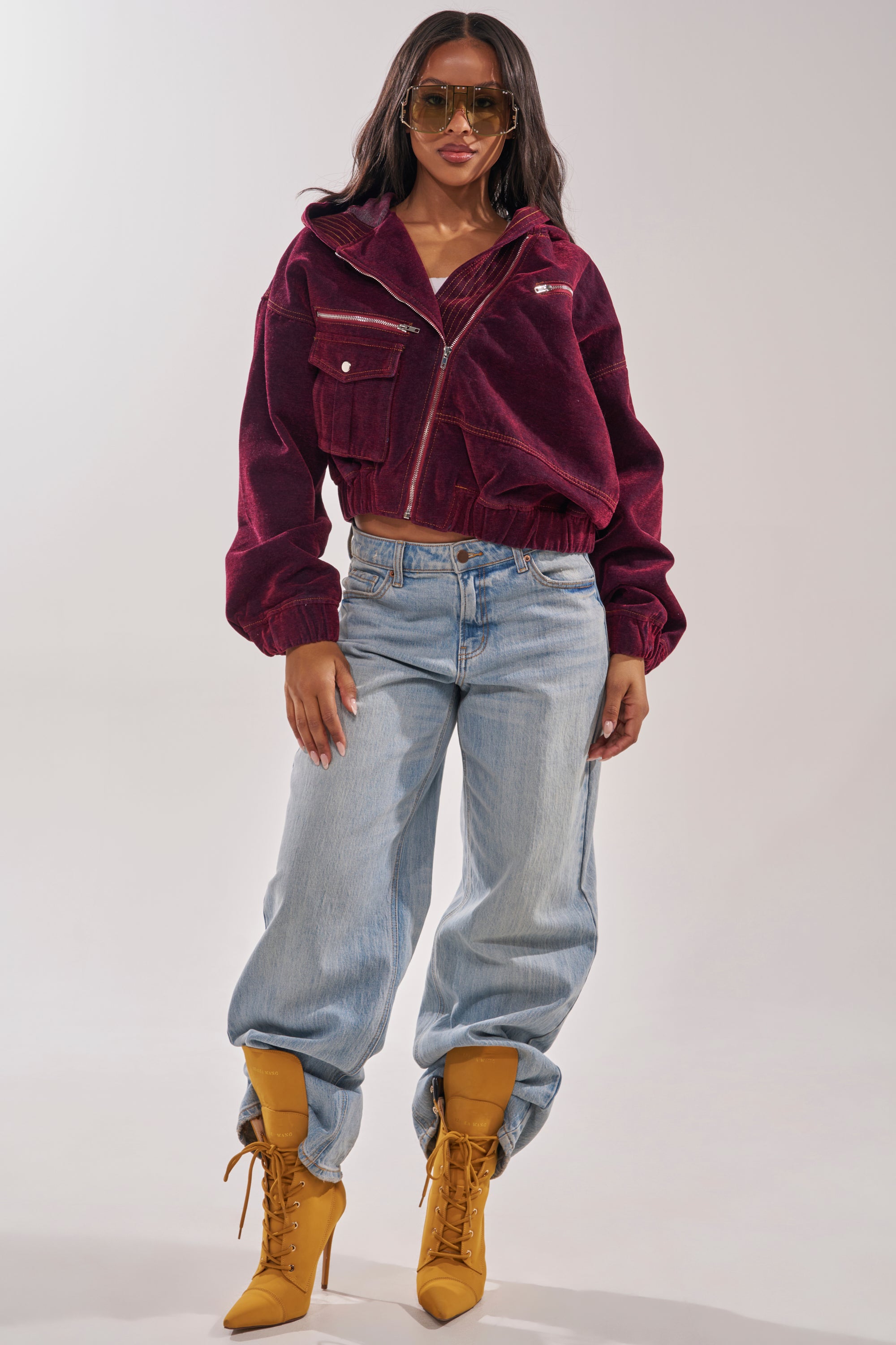 A woman stands confidently in the PAYTON VELVET DENIM BOMBER, pairing its maroon velvet and oversized hood with light blue baggy jeans, yellow lace-up heeled boots, and large sunglasses against a simple backdrop. 7