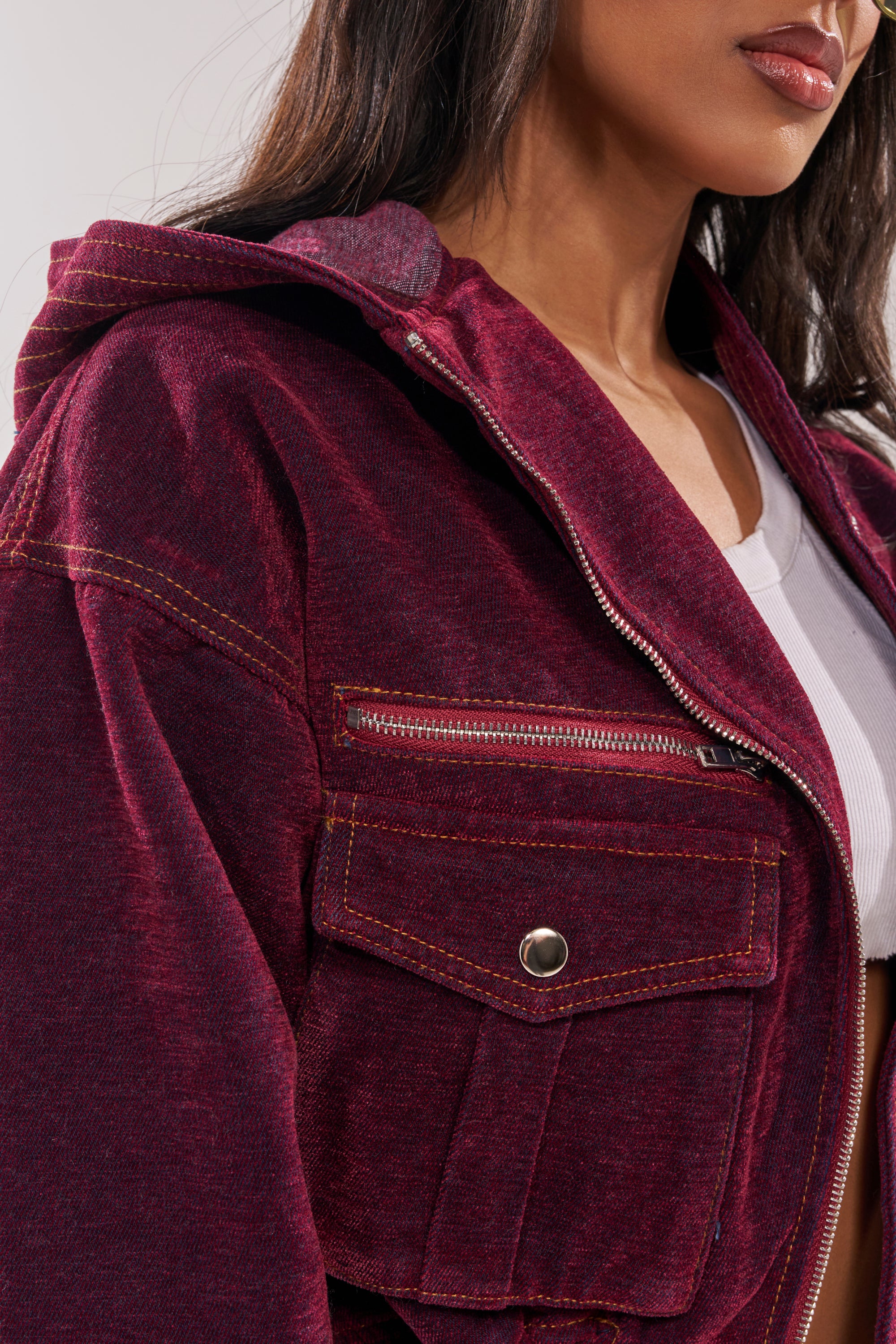 A close-up shows a woman in the PAYTON VELVET DENIM BOMBER, featuring an oversized hood and front pockets over a white top. Her face is partly visible, highlighting the bomber's rich texture, stitching, and modern streetwear style. 8