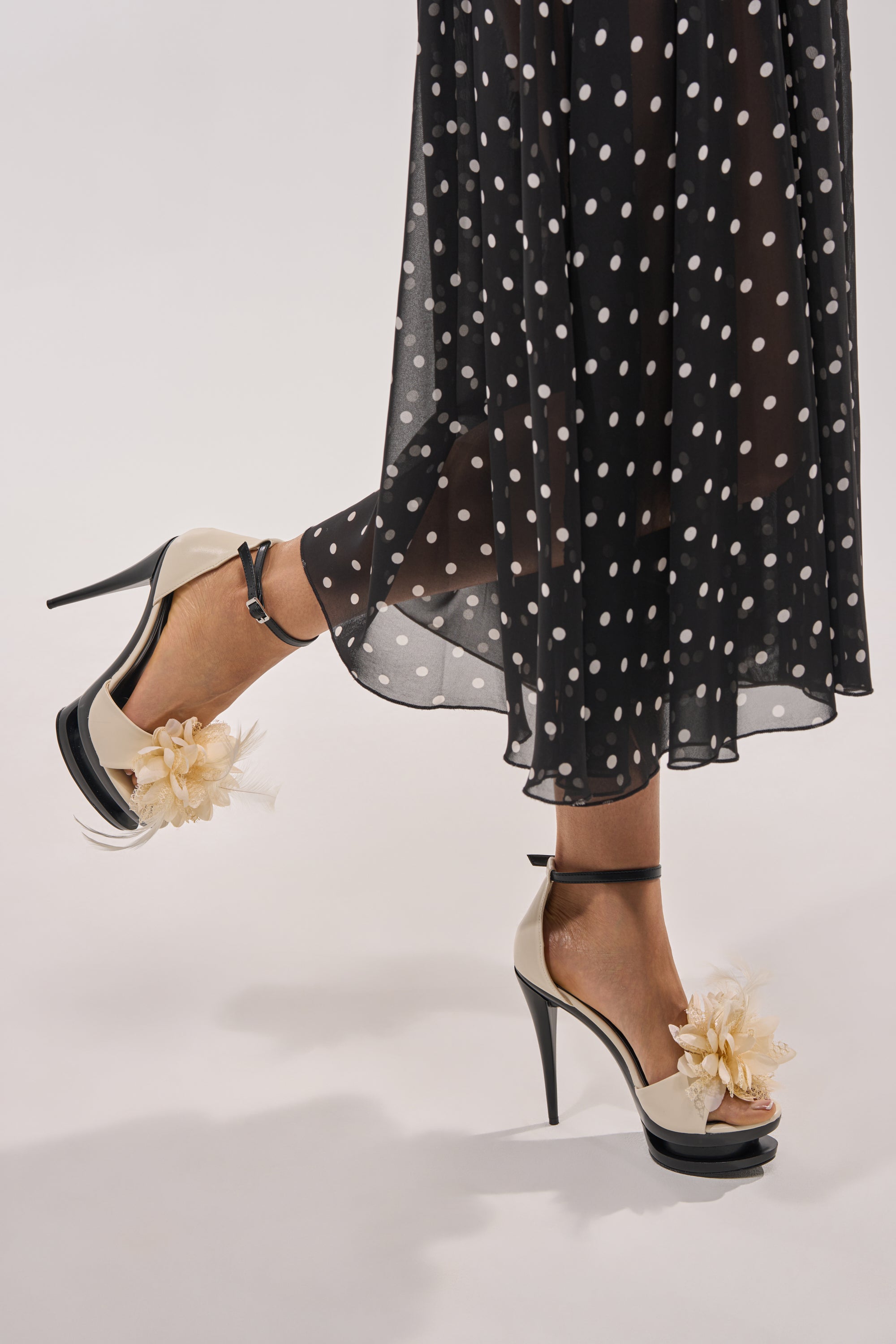 Wearing black and white polka dot sheer pants and AZALEA WANG VELVETINE IVORY STILETTO PLATFORM SANDALS, a person lifts one foot off the ground against a plain background. 2