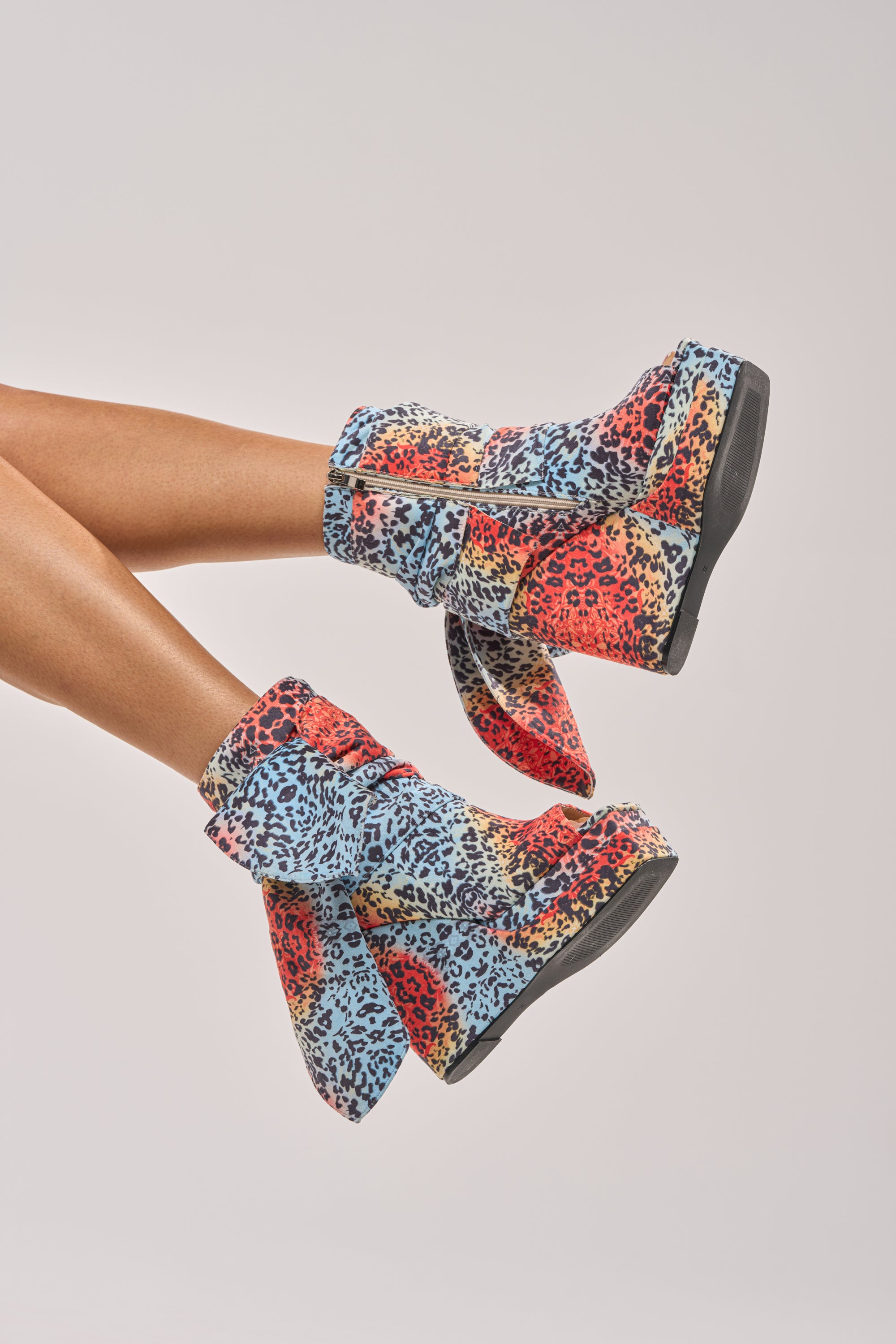 A pair of legs wearing AKIRA GOLD VENEZIO MULTI COLOR CHEETAH PRINT WEDGE BOOTIE with a colorful animal print and bold details, set against a plain light background. 0
