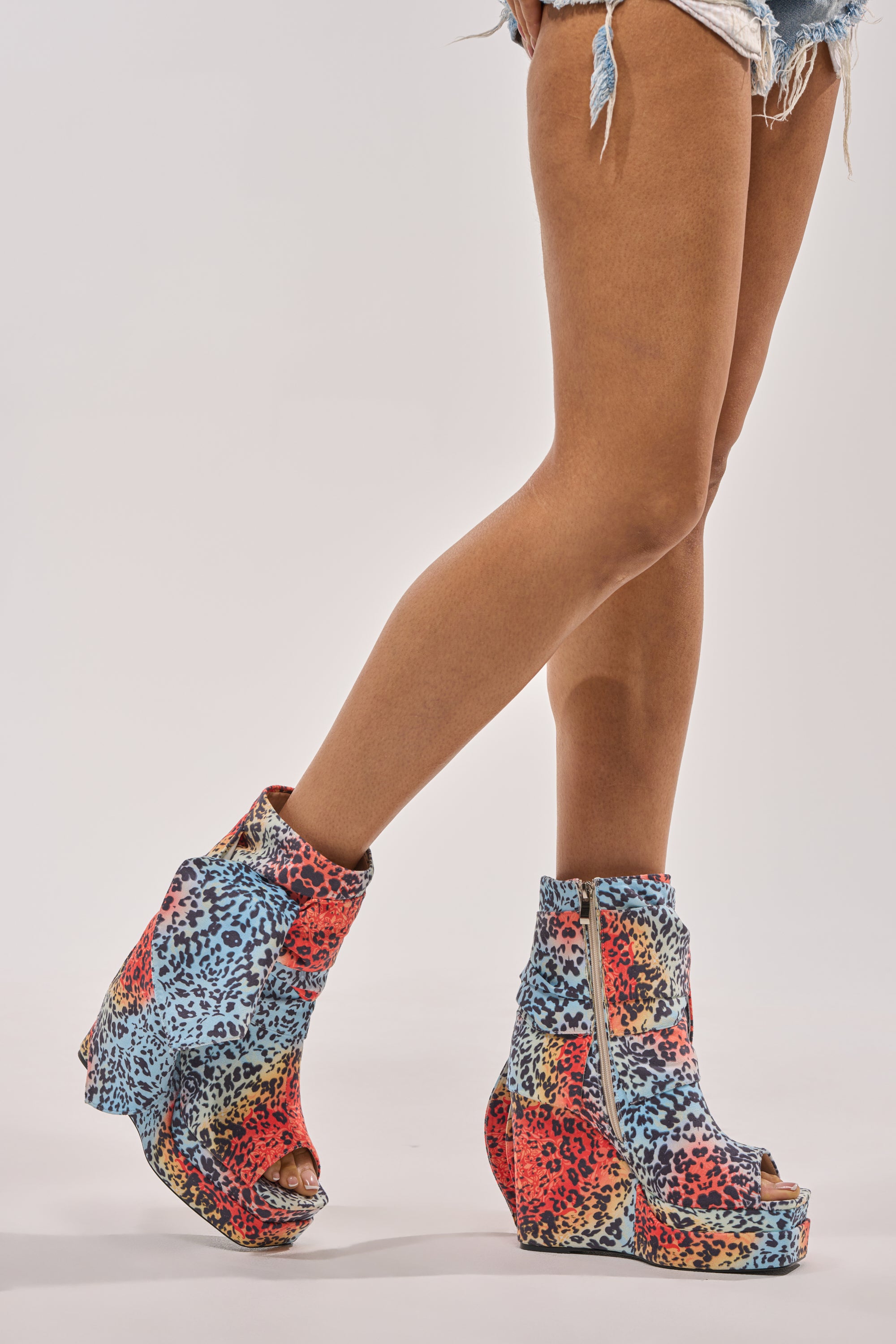 A person in distressed denim shorts wears the AKIRA Gold Venezio Multi Color Cheetah Print Wedge Bootie—platform, open-toe shoes with cheetah-print accents and front zippers—standing against a plain white background. 2