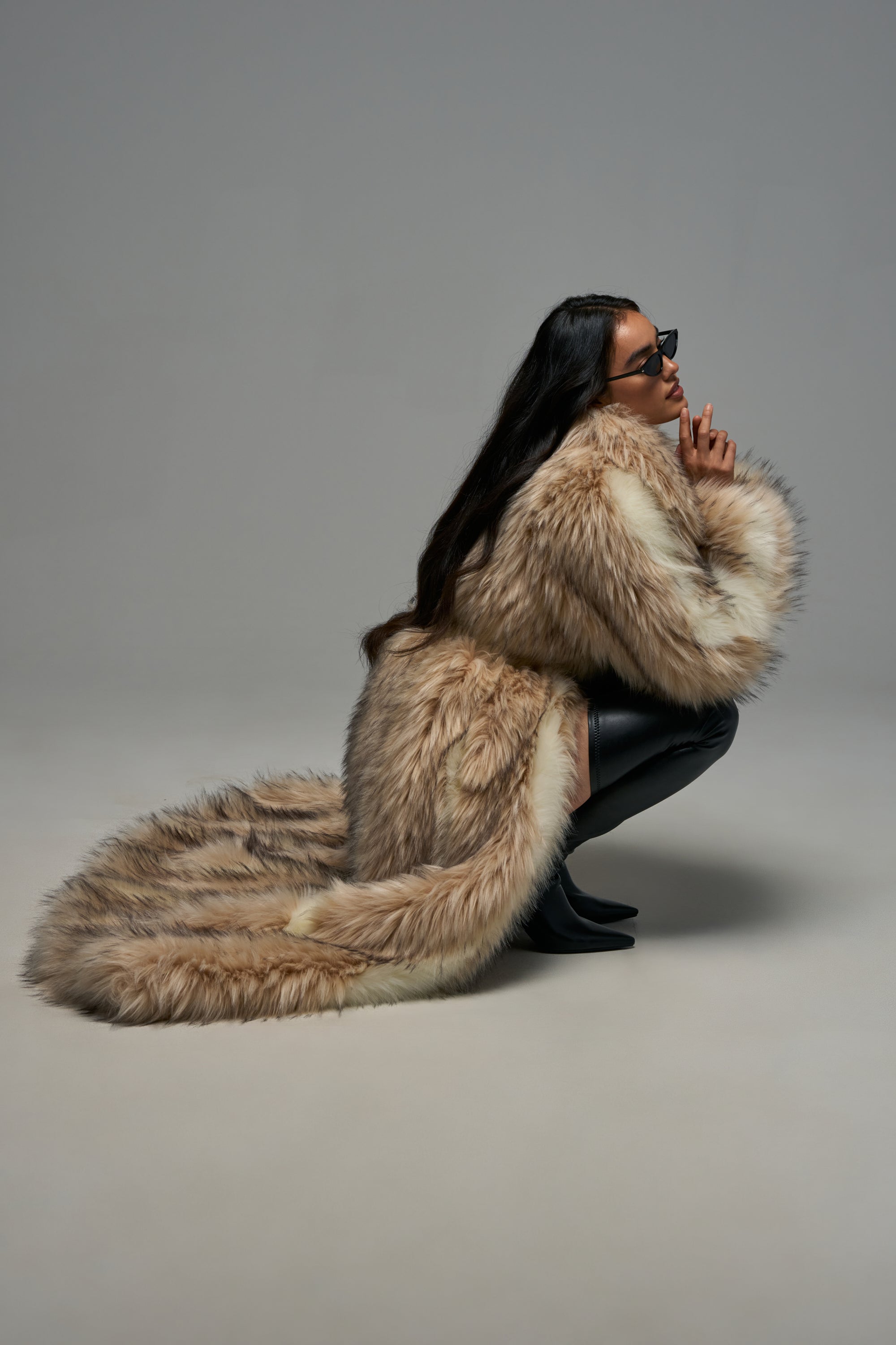 Wearing the VENUS FAUX FUR TRENCH—an oversized, statement winter coat that pools luxuriously on the floor—a person in sunglasses squats against a plain gray background, styled with black pants and shoes for a look that epitomizes luxurious cold-weather fashion.