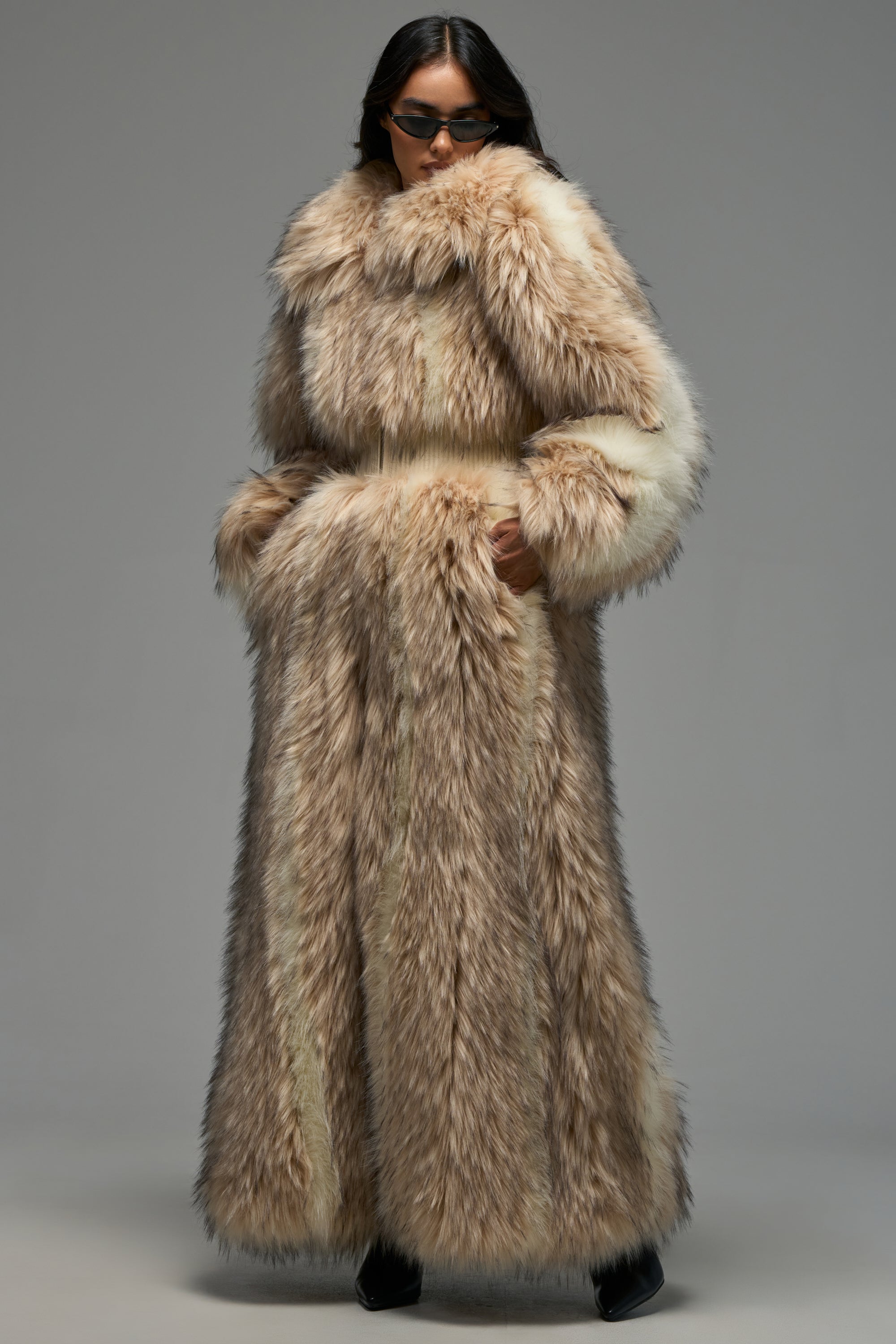 Wearing the VENUS FAUX FUR TRENCH—a long, thick, light brown statement winter coat—a person with dark sunglasses stands with their hands in the coat pockets against a plain gray background.