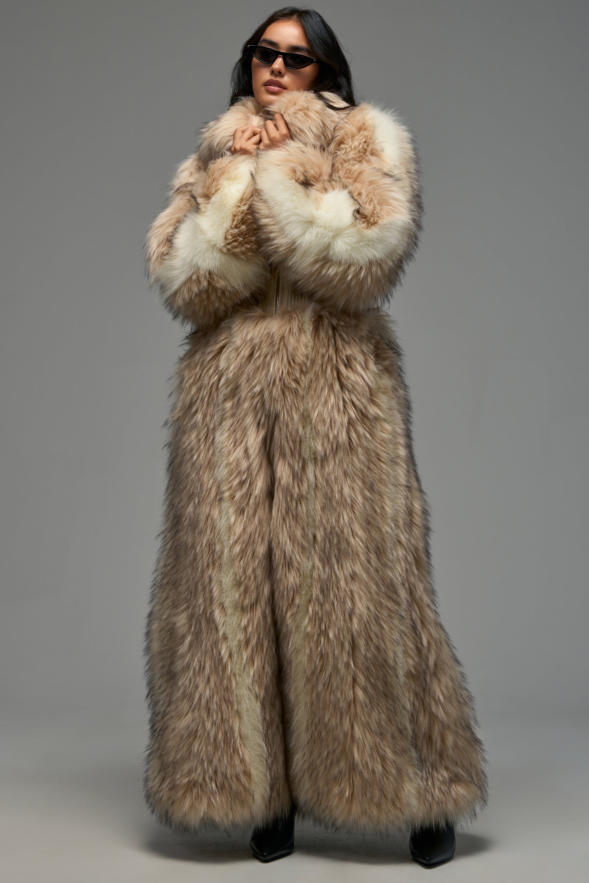 Wearing the VENUS FAUX FUR TRENCH—an oversized, long, thick coat in brown and white—a person stands against a plain background with dark sunglasses and hands held at their collar, perfectly exemplifying luxurious cold-weather style.