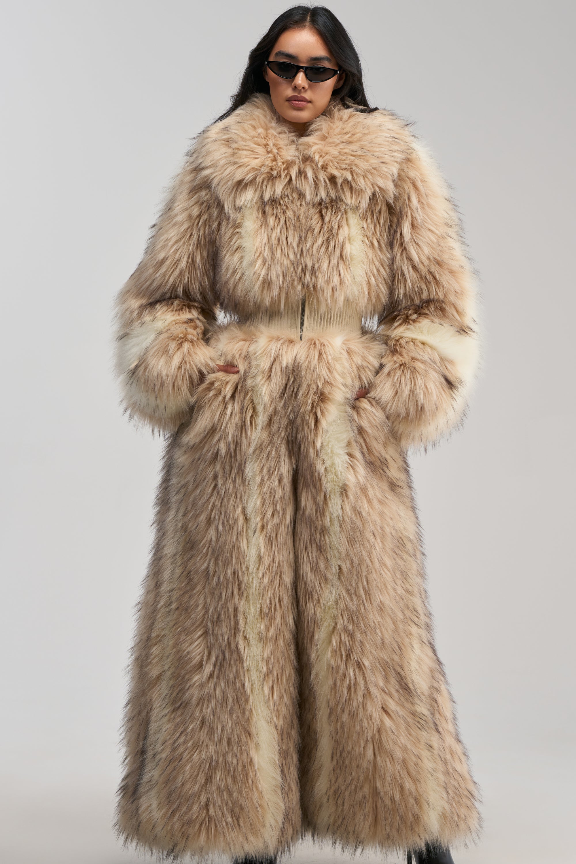 A person wearing sunglasses and the VENUS FAUX FUR TRENCH—a long, luxurious statement winter coat in tan and cream with wide sleeves—stands confidently against a plain background. The oversized design of the VENUS FAUX FUR TRENCH exudes sophisticated, cold-weather styling.