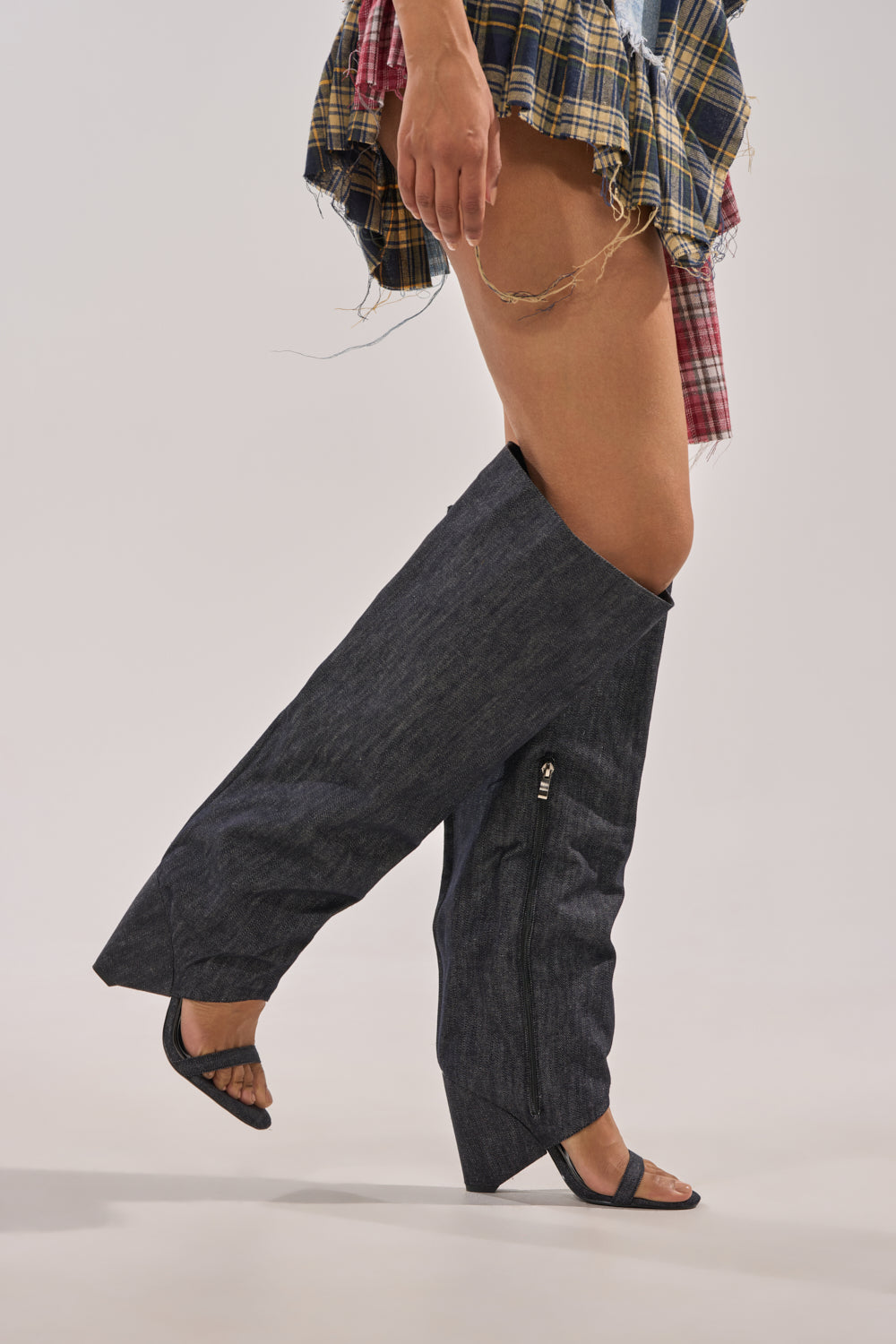 A person wears oversized denim pants with a frayed, asymmetrical plaid skirt, styled with the AZALEA WANG VEONA DARK DENIM OVER THE KNEE CHUNKY HEEL SANDAL. The background is plain and light-colored. 1