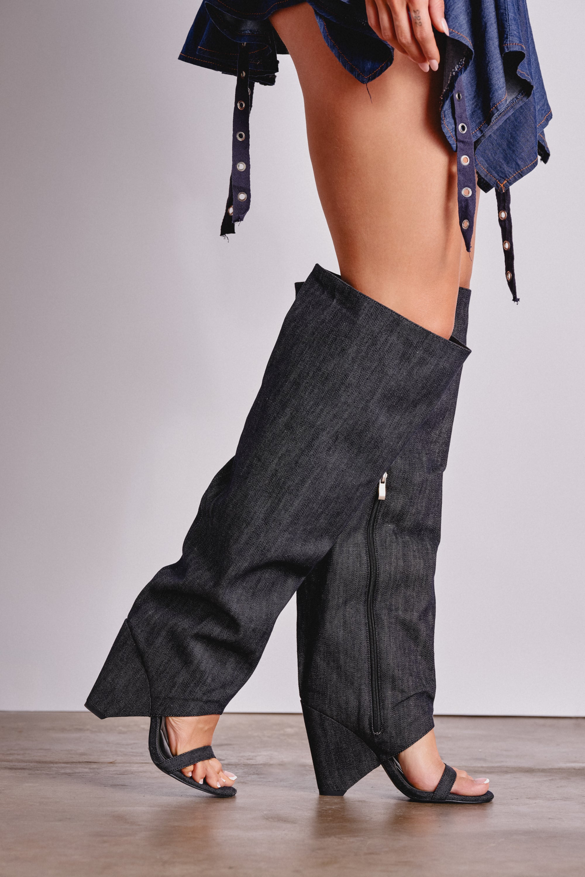 A person wearing the AZALEA WANG VEONA DARK DENIM OVER THE KNEE CHUNKY HEEL SANDAL and open-toe black heeled sandals stands on a concrete floor, with only their lower half visible. 0