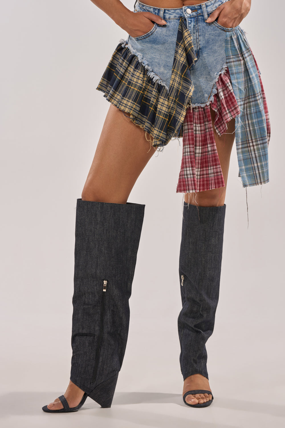 Wearing a mini skirt with denim and plaid patches, paired with the AZALEA WANG VEONA DARK DENIM OVER THE KNEE CHUNKY HEEL SANDAL, a person stands against a plain light background. 2