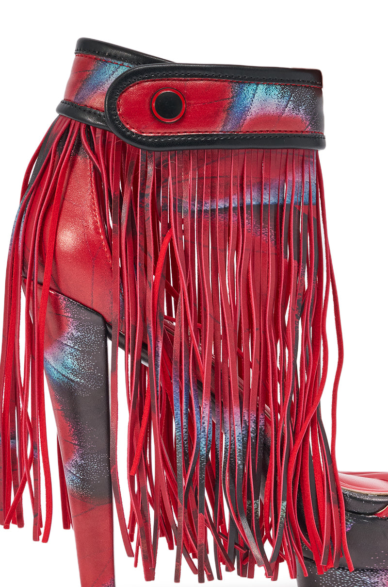 A close-up of the AKIRA GOLD PRINTED PU FRINGE STILETTO SANDAL with a colorful abstract platform heel, long red fringe, and a strap accented by a black button. 6