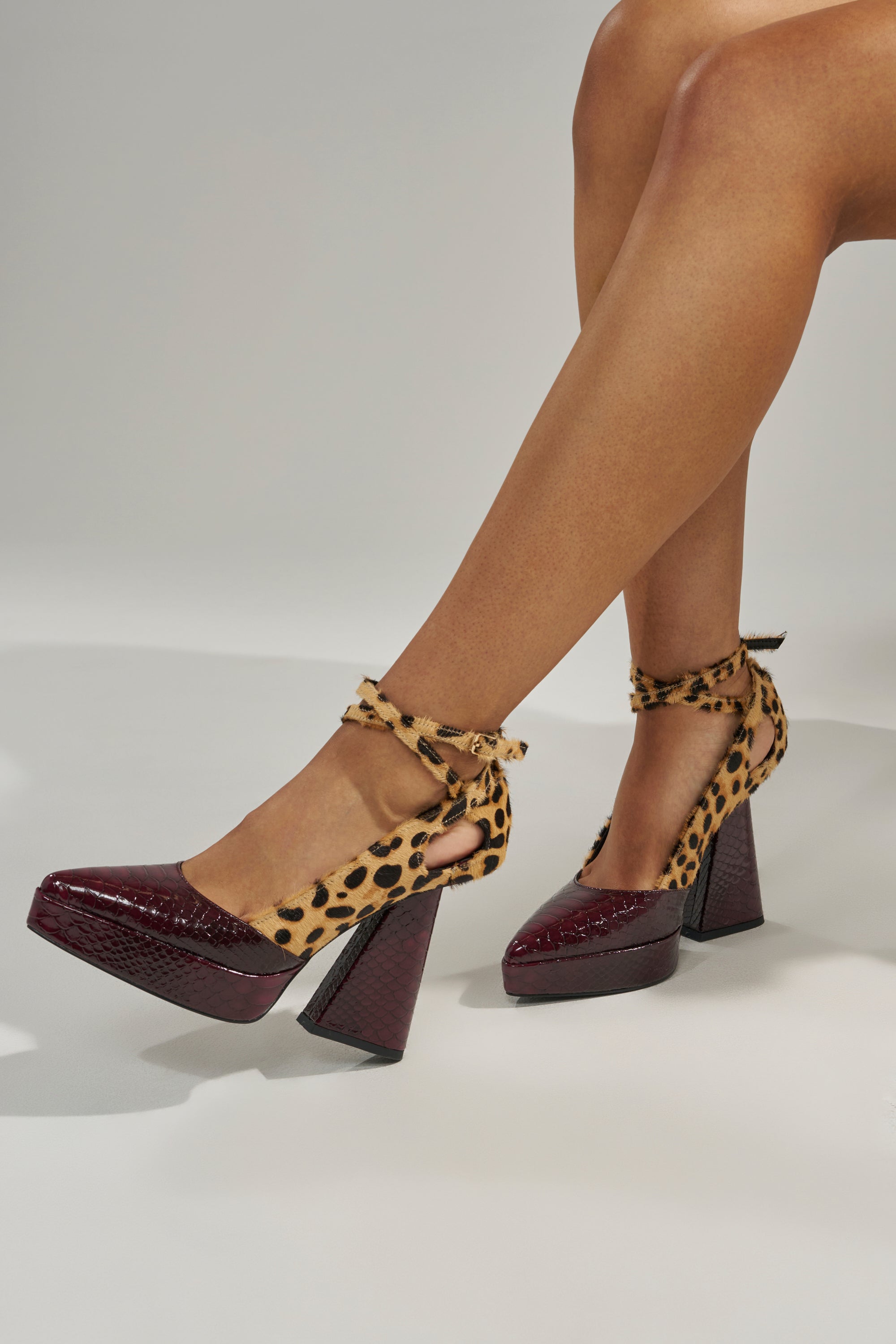 Someone wearing the AZALEA WANG VEVELYN MULTI CHUNKY PUMP, featuring a pointed toe, burgundy crocodile-patterned toes and heels, and leopard-print ankle straps and sides, is sitting with legs crossed.