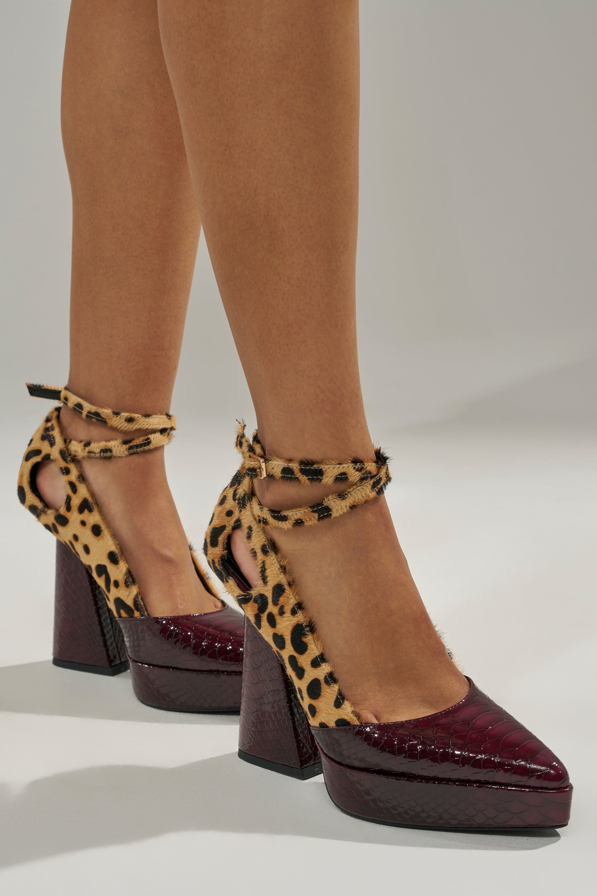 A pair of women's feet in AZALEA WANG VEVELYN MULTI CHUNKY PUMP shoes with burgundy, crocodile-textured toes, platform heels, and leopard-print ankle-wrap straps.