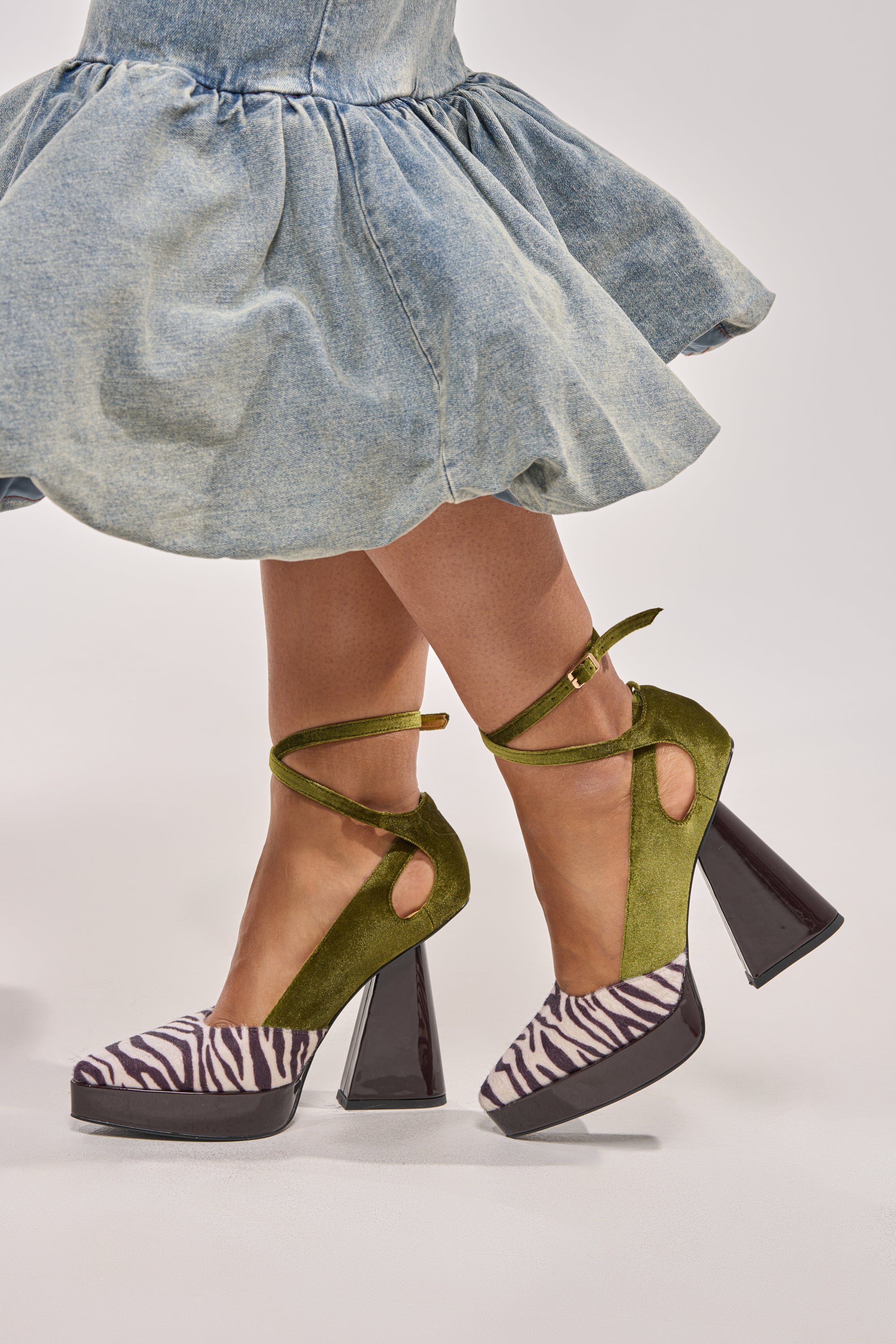 A person in a light blue ruffled skirt and zebra print platform heels stands against a plain background, showcasing the AZALEA WANG VEVELYN OLIVE BLOCK PUMP with velvet, chunky black block heels, and ankle straps. 1