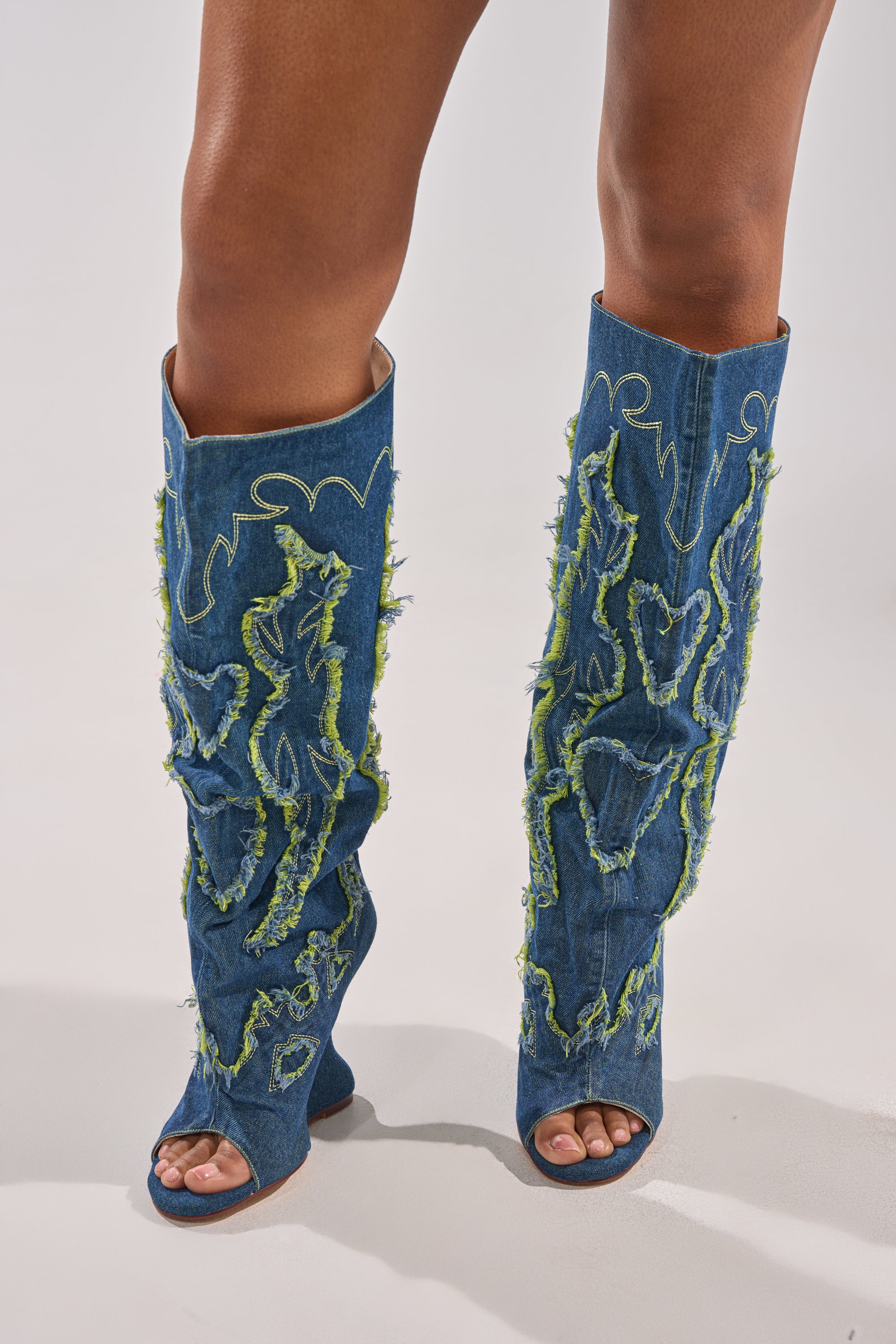 Only the lower legs and feet are visible as a person stands on a white surface, wearing the AZALEA WANG VIANCA GREEN MULTI DENIM WEDGE BOOT—knee-high denim boots with frayed green flame-like designs for bold Western style.
