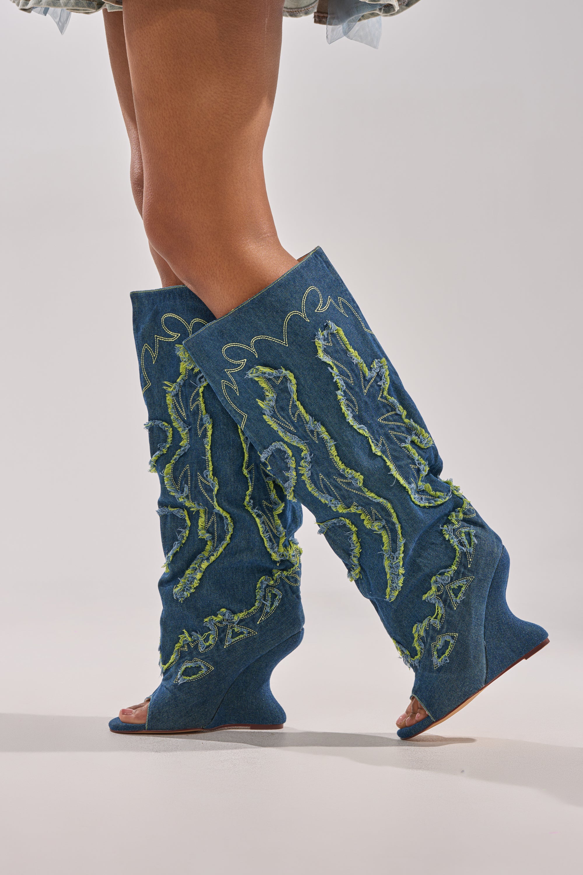 A close-up shows crossed legs wearing the AZALEA WANG VIANCA GREEN MULTI DENIM WEDGE BOOT, featuring knee-high denim with green textured designs, open toes, wavy wedge heels, and frayed embellishments against a light background.