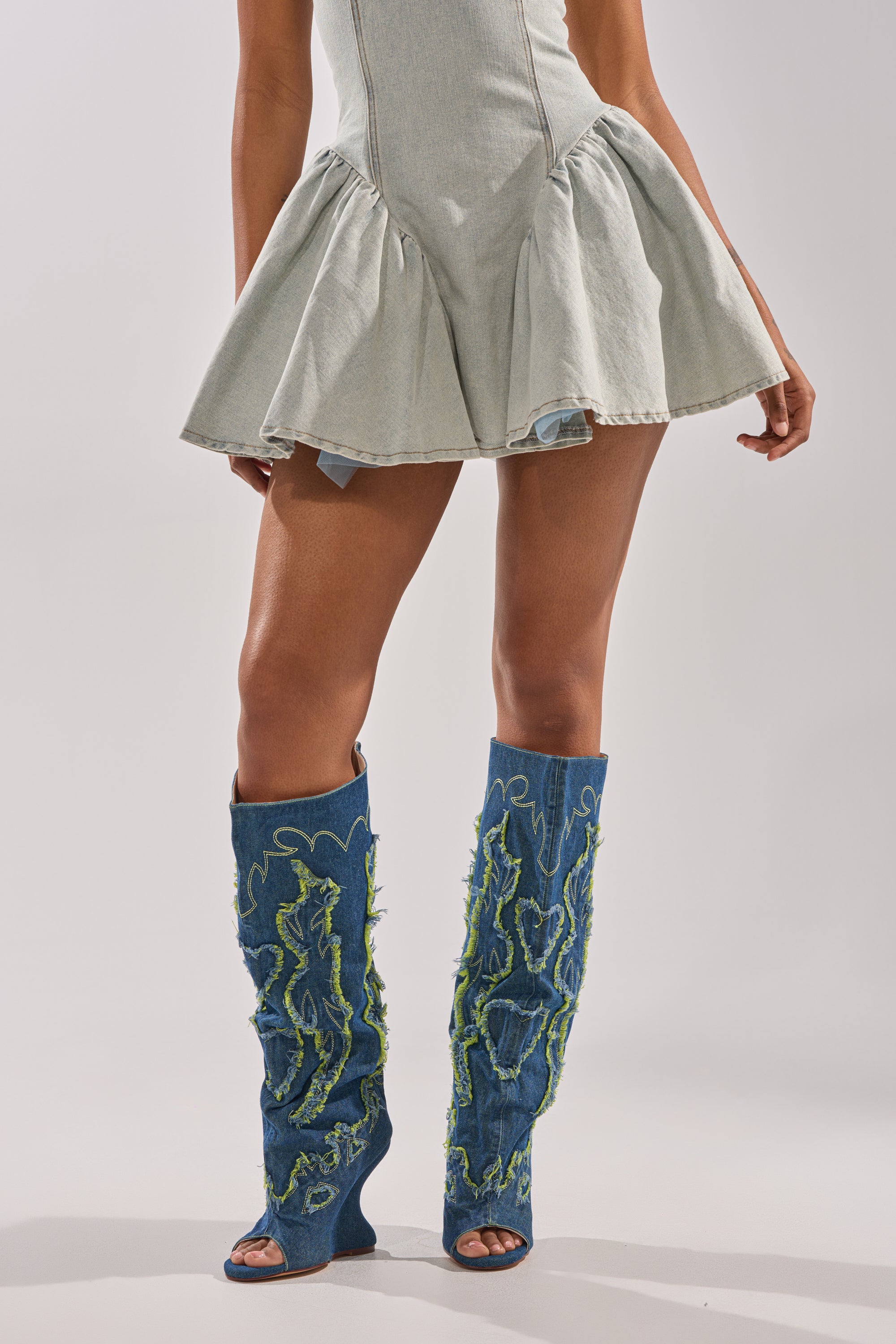 A person wears a light denim mini dress with a flared skirt and bold AZALEA WANG VIANCA GREEN MULTI DENIM WEDGE BOOT, featuring green textured designs, against a plain light background.
