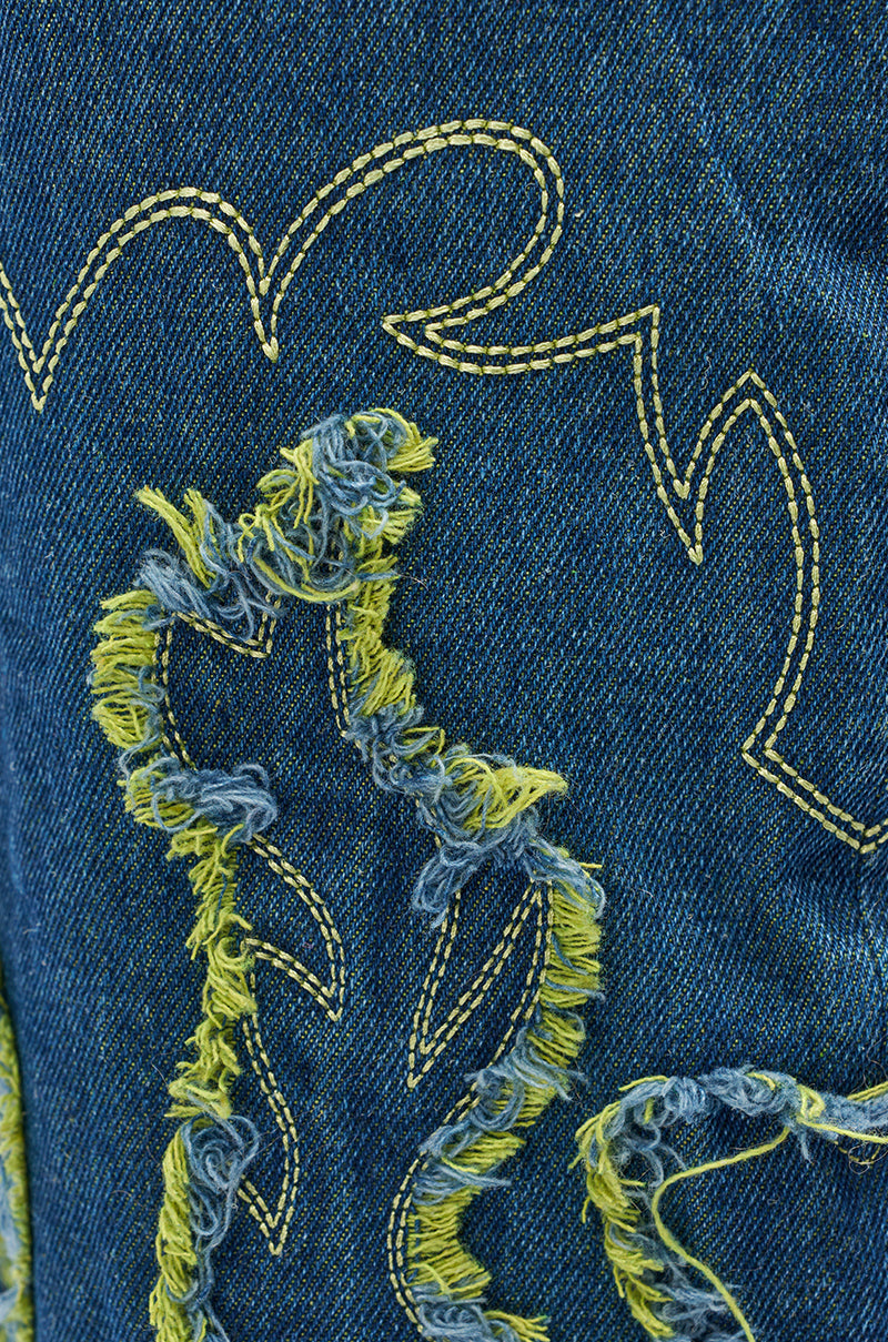 Close-up of the AZALEA WANG VIANCA GREEN MULTI DENIM WEDGE BOOT's blue denim fabric, featuring yellow and white frayed embroidery in abstract shapes, echoing classic Western boot stitching and a distressed textured look.