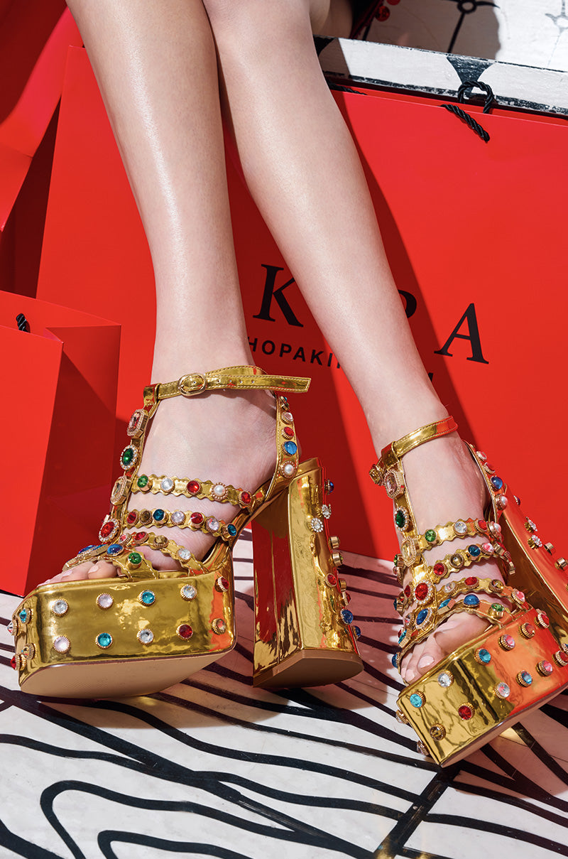 A close-up shows a person flaunting the AZALEA WANG Vianni Gold Chunky Sandals, adorned with multicolor gemstone embellishments. Paired with red shopping bags in the background, the scene bursts with vibrancy and style, while comfort cushion insoles ensure every step is taken in stylish comfort.
