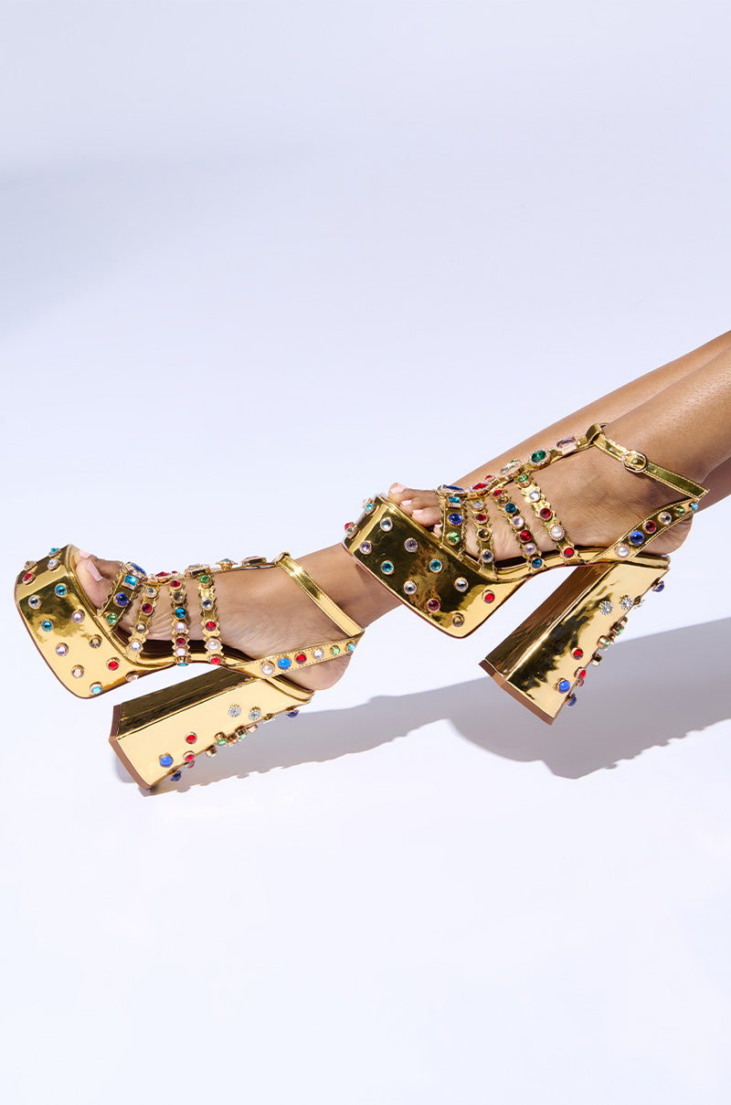 The AZALEA WANG VIANNI GOLD CHUNKY SANDAL, adorned with multicolor gemstone embellishments, draws attention with its bold design. Featuring thick high heels and multiple straps, these dazzling sandals stand out against the plain white background. Enhanced by a comfort cushion insole, they offer effortless elegance all day long.