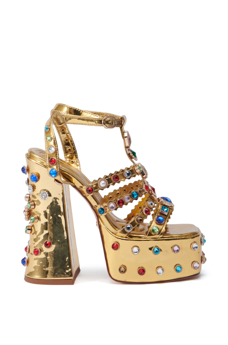 The AZALEA WANG VIANNI Gold Chunky Sandal showcases a luxurious design with its platform high heels adorned with multicolor gemstone embellishments and an ankle strap. Featuring a comfort cushion insole, these sandals blend style and comfort beautifully, highlighted by vibrant red, blue, and clear stones on a textured surface.