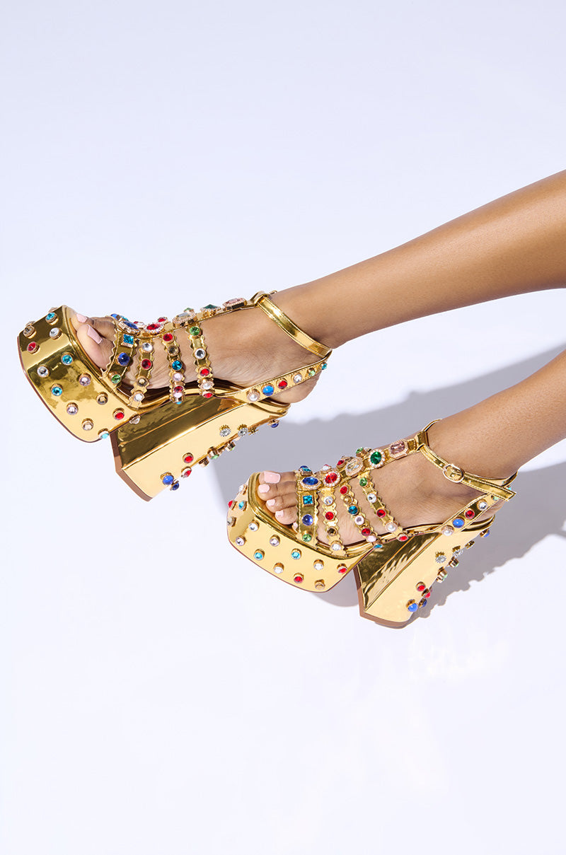 The AZALEA WANG VIANNI GOLD CHUNKY SANDAL is adorned with multicolor gemstones, featuring elegant ankle straps and high heels, beautifully showcased against a pristine white backdrop.