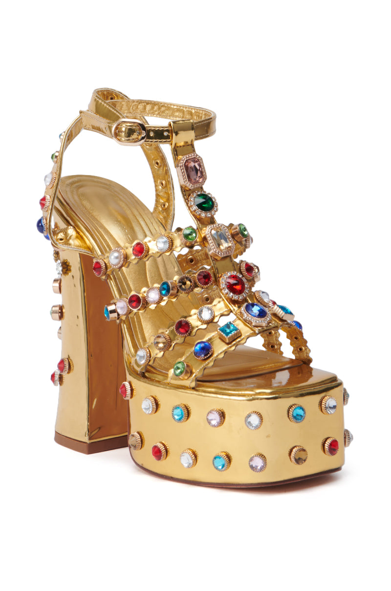 The AZALEA WANG VIANNI GOLD CHUNKY SANDAL is a bold, glamorous design featuring a comfortable cushion insole and adorned with multicolor gemstone embellishments in shades of red, blue, and green. It also includes an ankle strap and a thick heel.