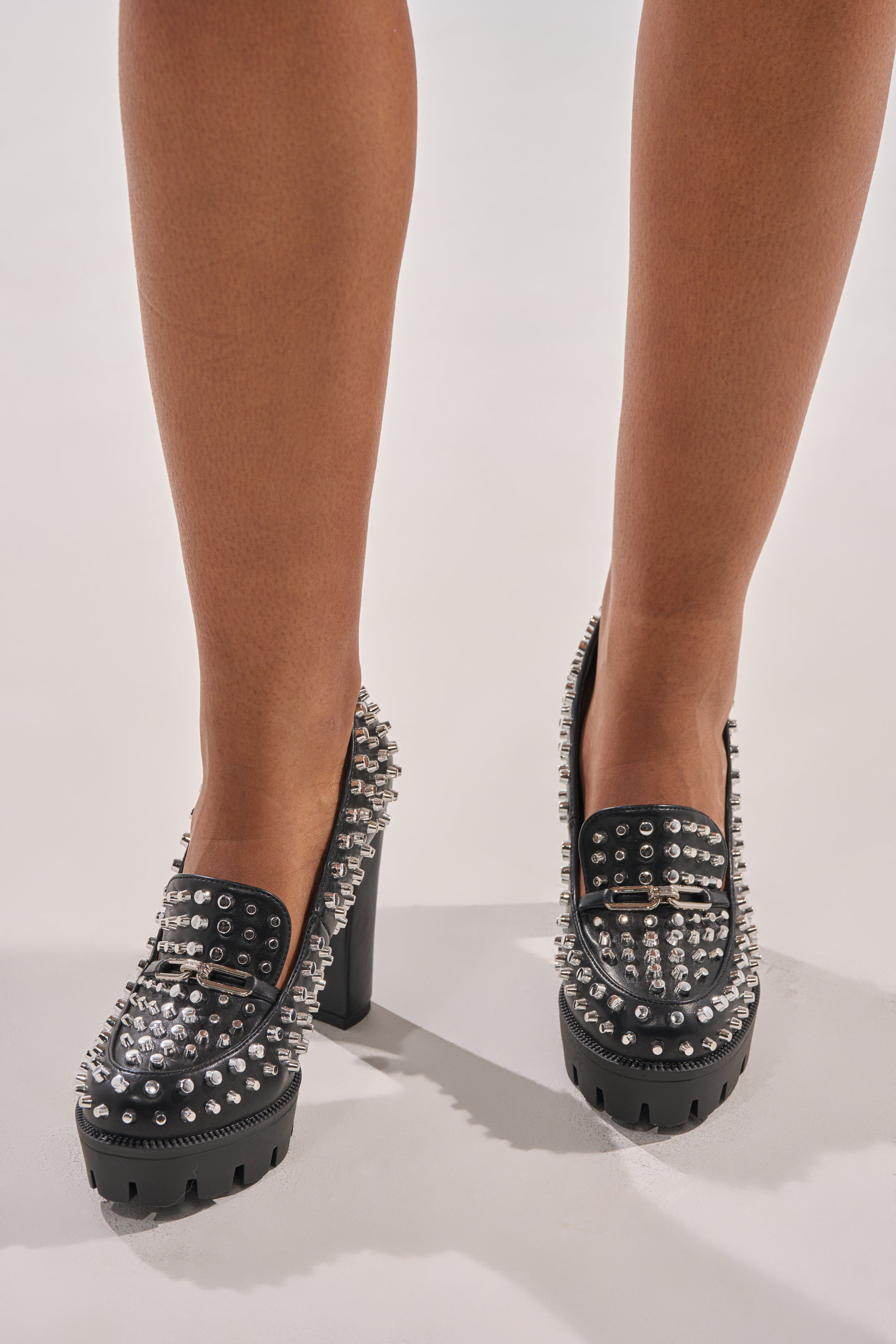 A person wearing the AKIRA GOLD VICTOIRE BLACK STUDDED PUMP LOAFER, featuring black faux leather, silver studs, chunky platform heels, and high soles, is standing on a light-colored surface. 1