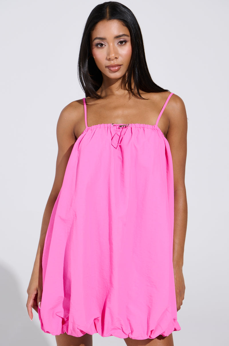 A person with long dark hair wears a bright pink REBECCA BUBBLE MINI DRESS from the AKIRA Label. The dress features thin straps, a gathered neckline, and a voluminous hem as they pose against a plain, light-colored background.
