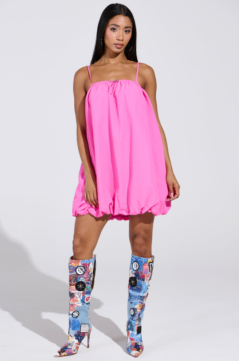 A woman wears a bright pink REBECCA BUBBLE MINI DRESS with thin straps, pairing it with colorful graphic knee-high boots. The simple, light background highlights the AKIRA Label-inspired ensemble.