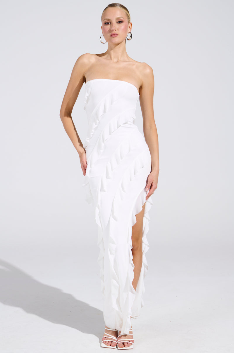 A person confidently poses against a plain backdrop in the ELLA MESH TUBE DRESS IN WHITE with elegant ruffle detailing. They're accessorized with hoop earrings and open-toed heels.
